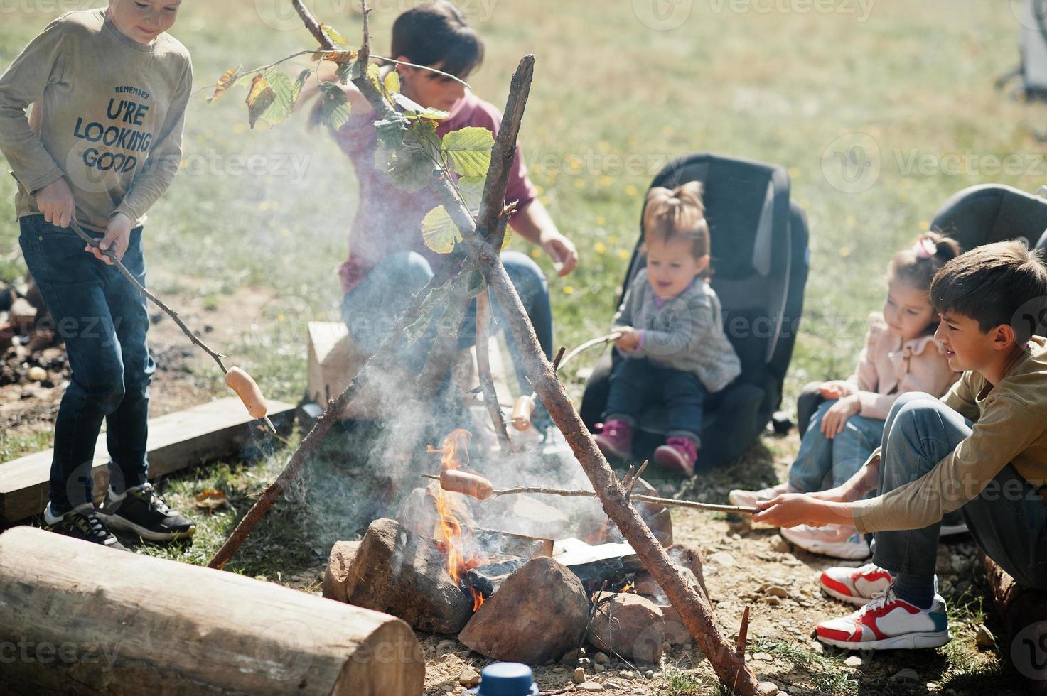 Familienfeuer im Berg. Bratwürste. Mutter mit vier Kindern Camping. Herbstwanderung und Lagerwetter. Erwärmen und Kochen in der Nähe einer Flamme zusammen. foto