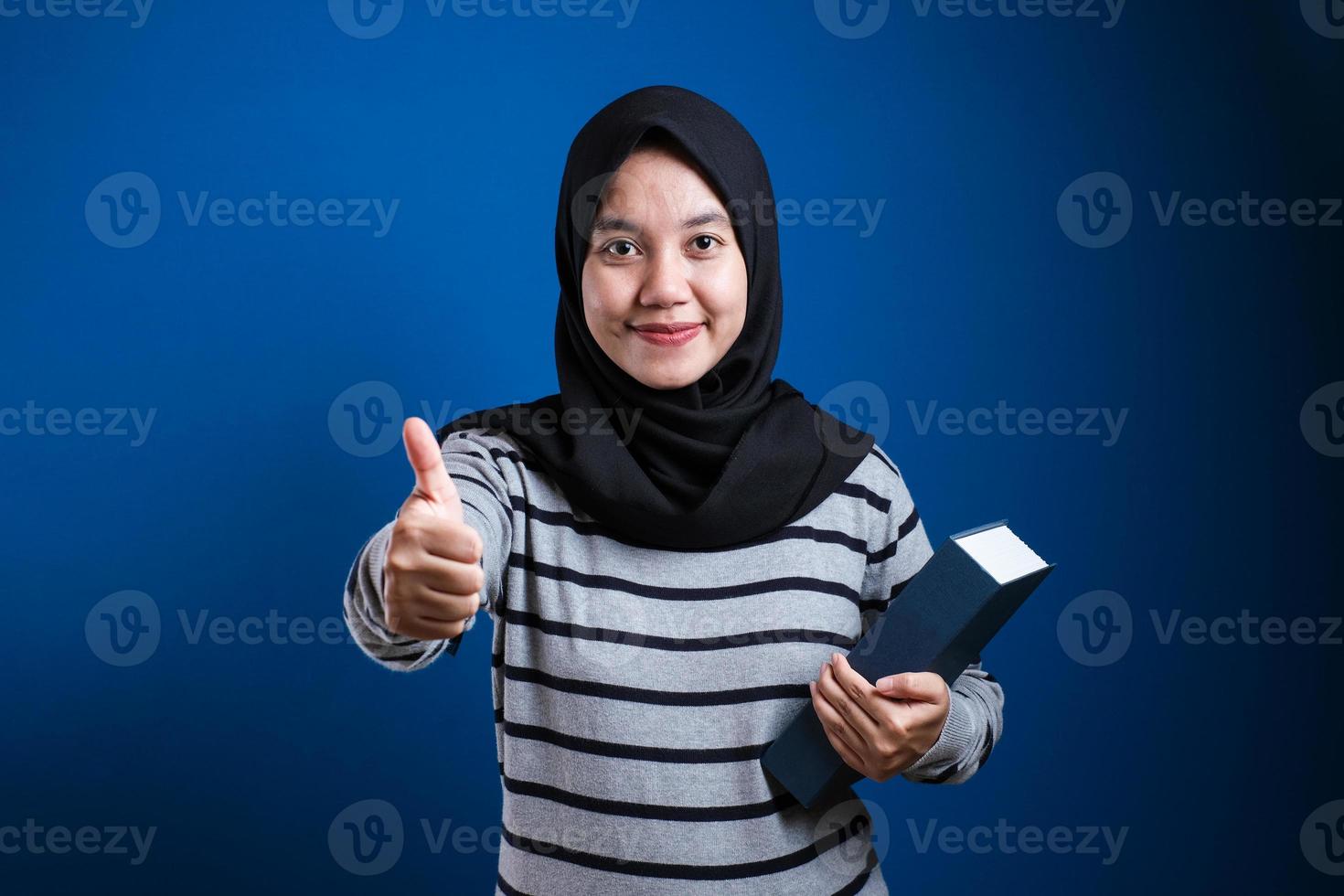 junge schöne studentin mit muslimischem hijab, die bücher hält und fröhliche daumen hoch geste mit der hand macht foto