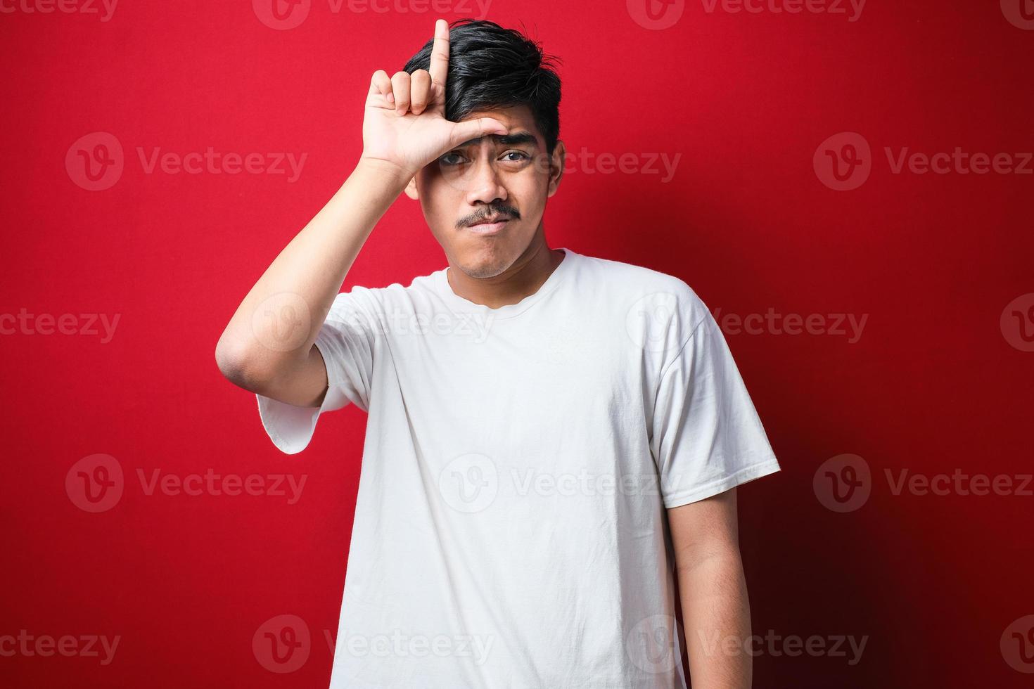 junger gutaussehender mann mit weißem t-shirt, der sich über menschen mit fingern auf der stirn lustig macht, die verlierergesten verspotten und beleidigen. foto