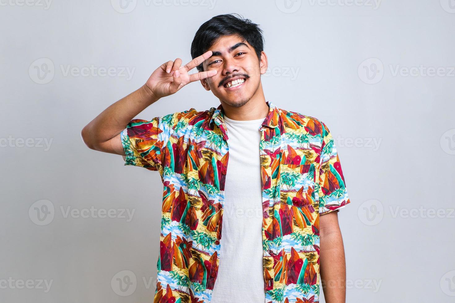 junger hübscher asiatischer kerl, der lässiges hemd trägt, das friedenssymbol mit den fingern über dem gesicht macht foto