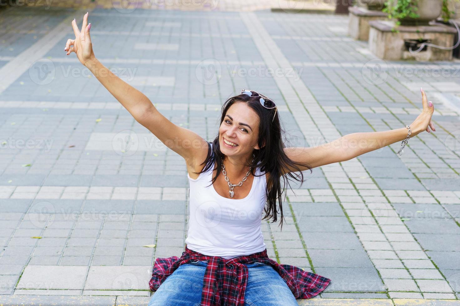 glückliche junge Modefrau, die auf dem stilvollen weiblichen Modell des Straßenbürgersteigs sitzt foto