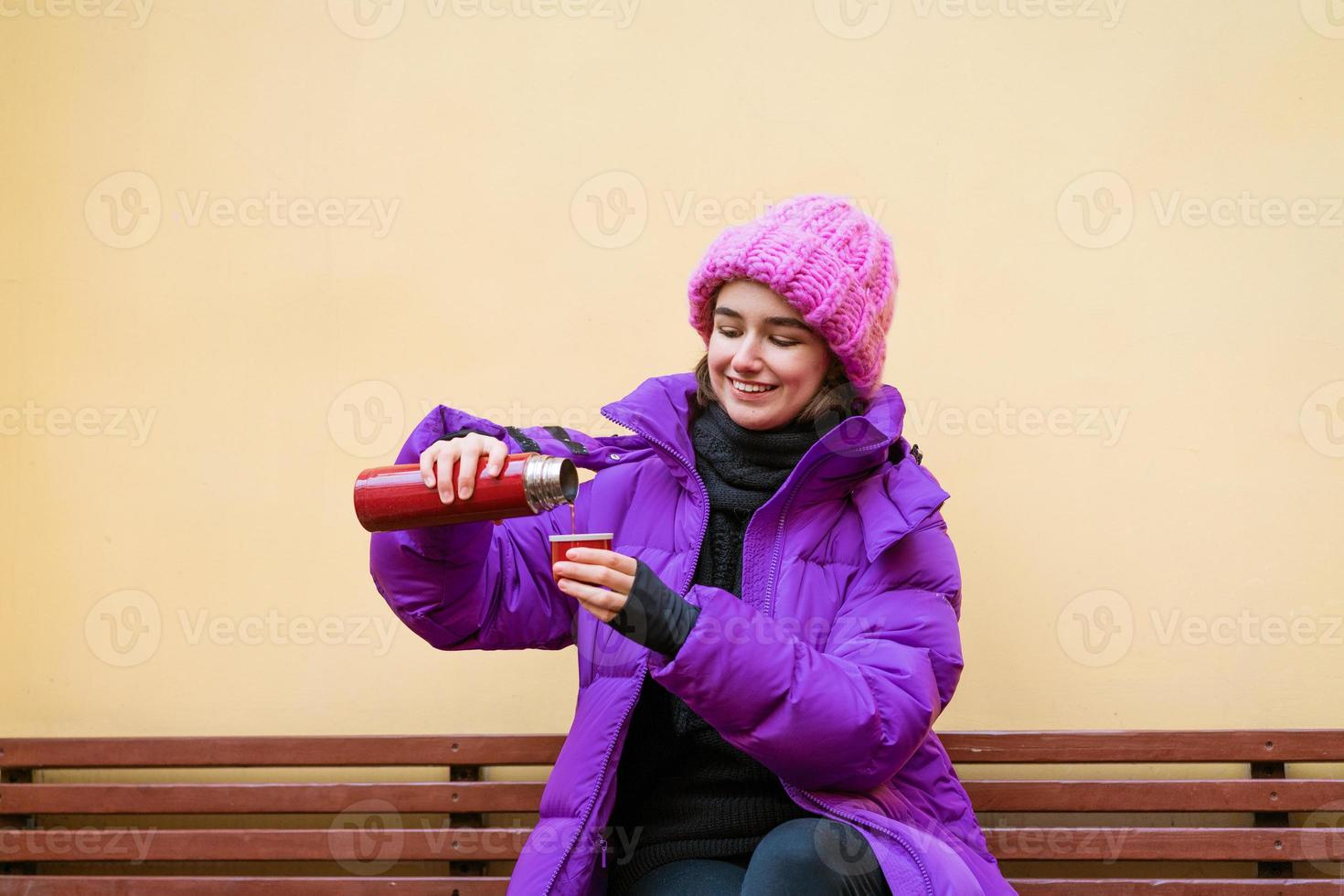 eine junge frau in warmer oberbekleidung gießt sich im sitzen heißen tee aus einer thermoskanne ein foto