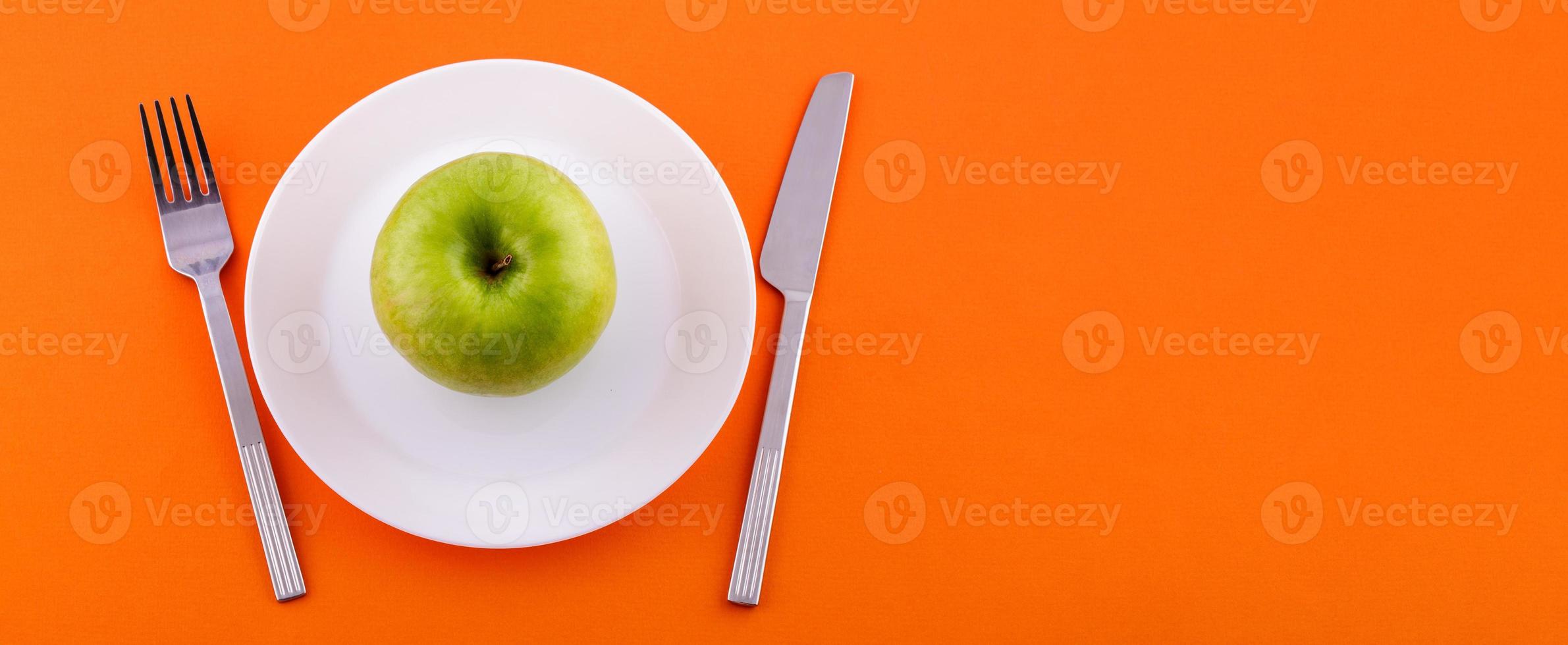 auf einem Teller ein grüner Apfel und ein Messer mit einer Gabel auf orangefarbenem Hintergrund foto