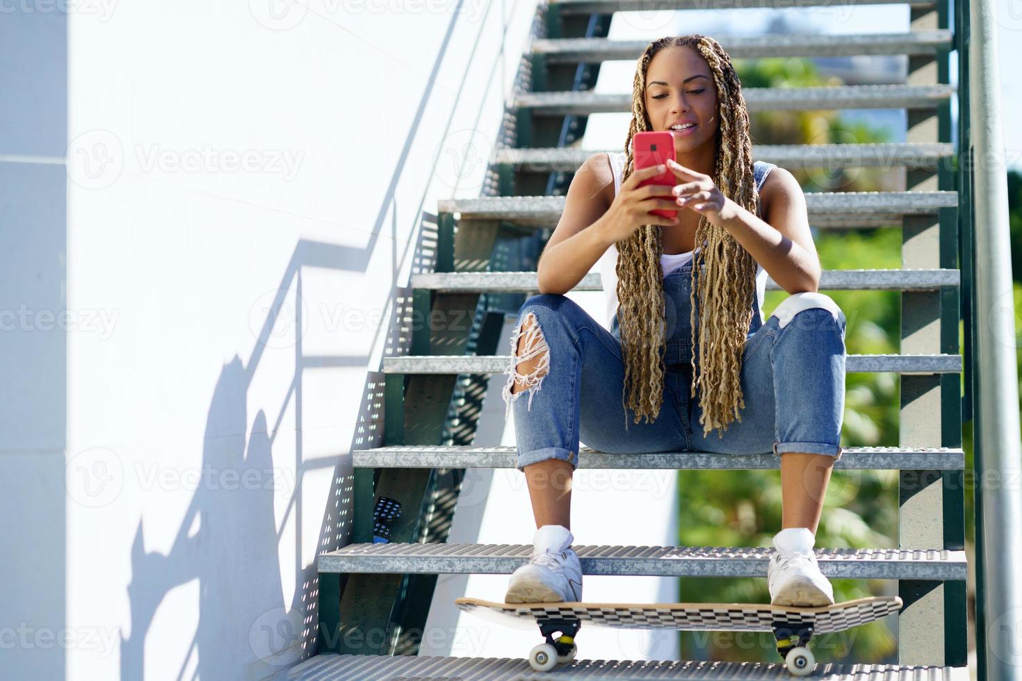 Schwarze Frau mit farbigen Zöpfen, die ihr Smartphone konsultiert, während ihre Füße auf einem Skateboard ruhen. foto