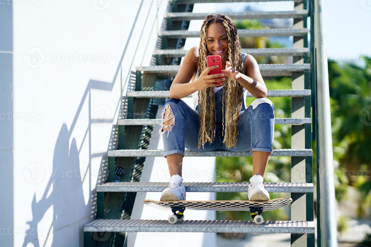 Schwarze Frau mit farbigen Zöpfen, die ihr Smartphone konsultiert, während ihre Füße auf einem Skateboard ruhen. foto