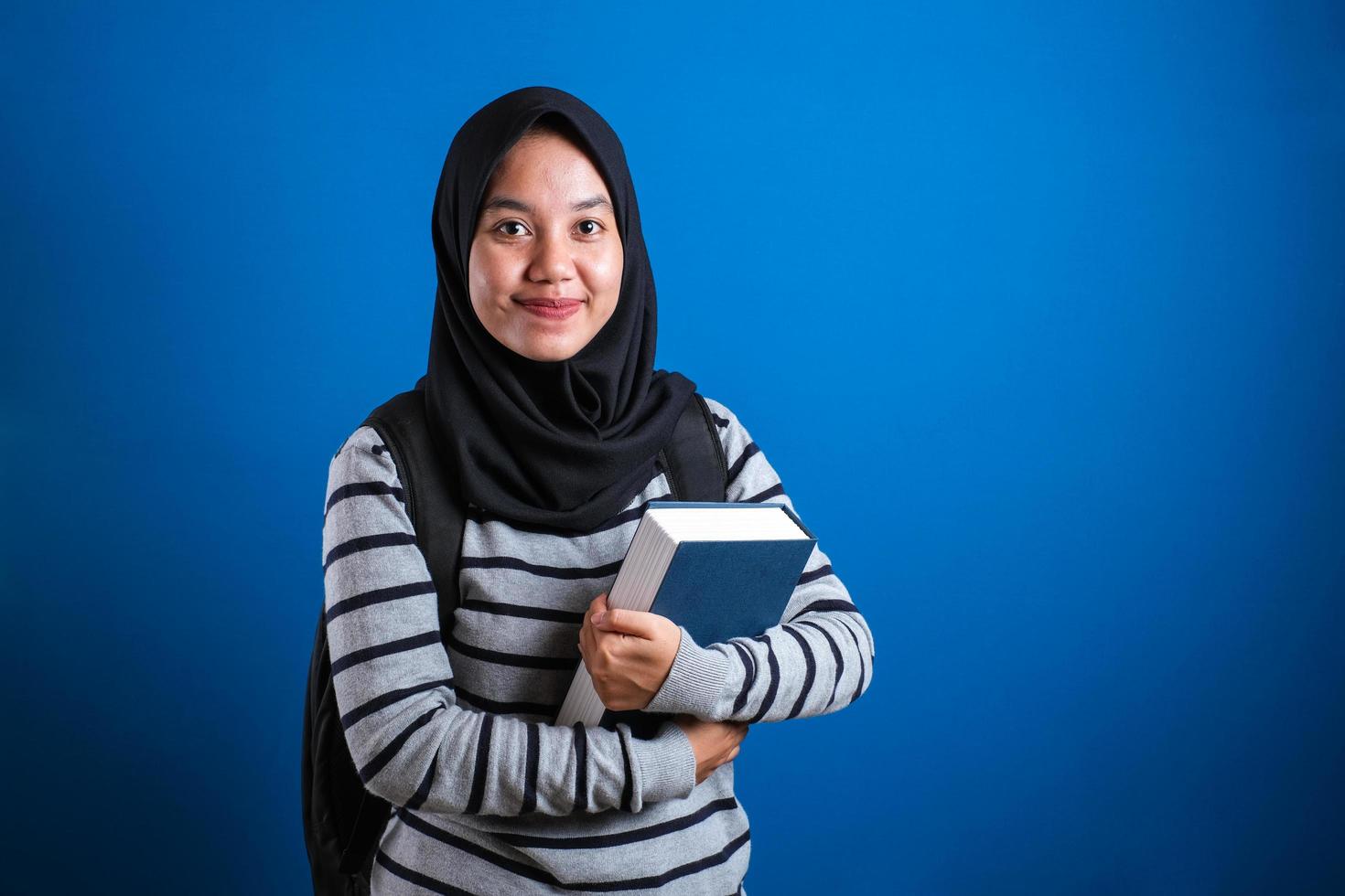 asiatisches muslimisches studentenmädchen, das hijab trägt, lächelt freundlich und hält großes buch foto