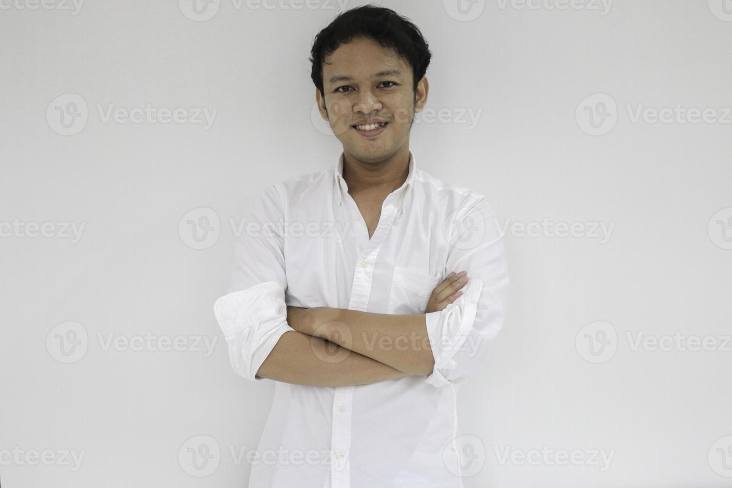 Der junge asiatische Mann mit weißem Hemd hält die Hände mit einem glücklichen Lächeln und Selbstvertrauen gekreuzt. erfolgreicher indonesischer mann auf grauem hintergrund. foto