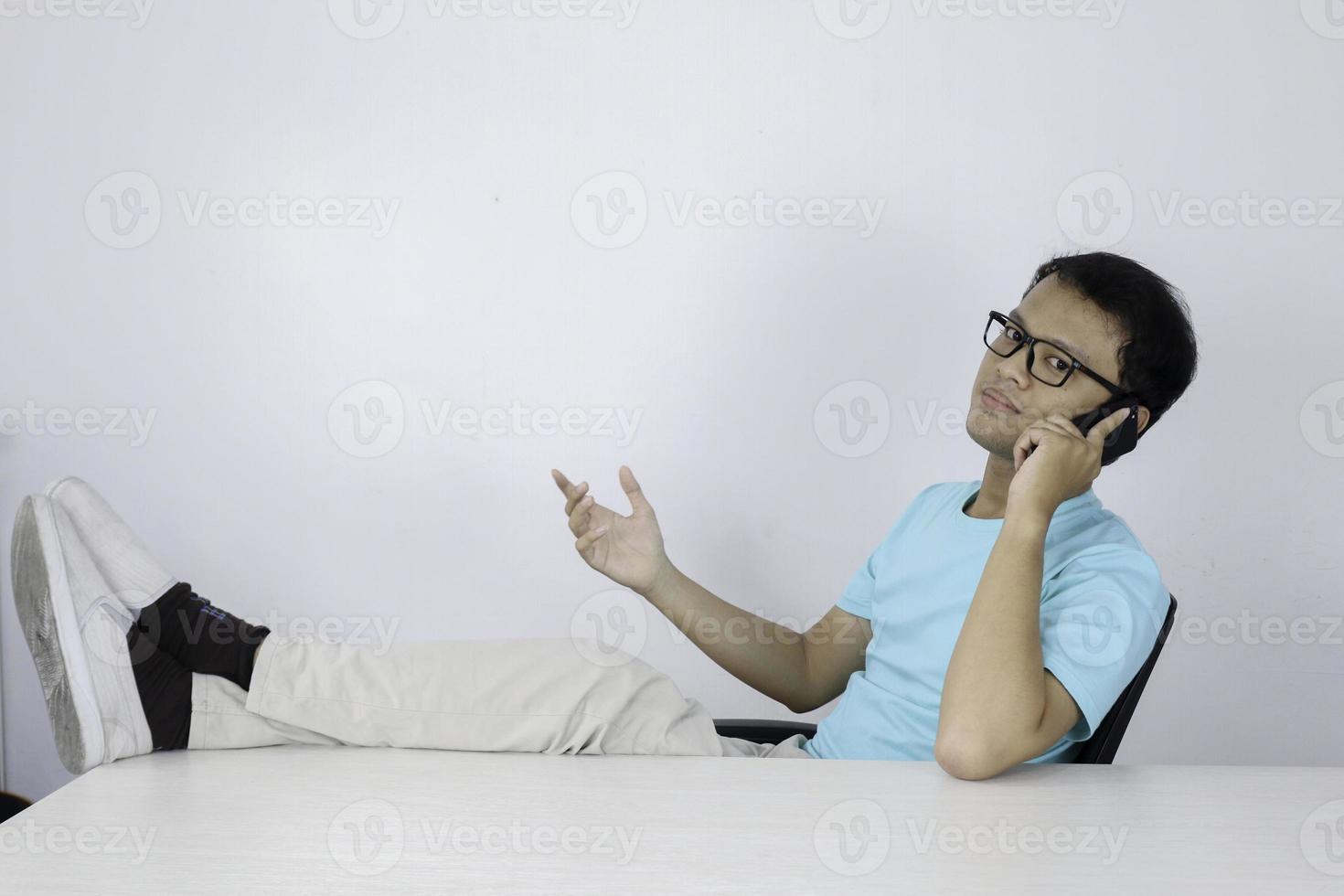 junger asiatischer mann fühlt sich glücklich und lächelt, wenn er mit dem bein auf dem tisch am telefon spricht. foto