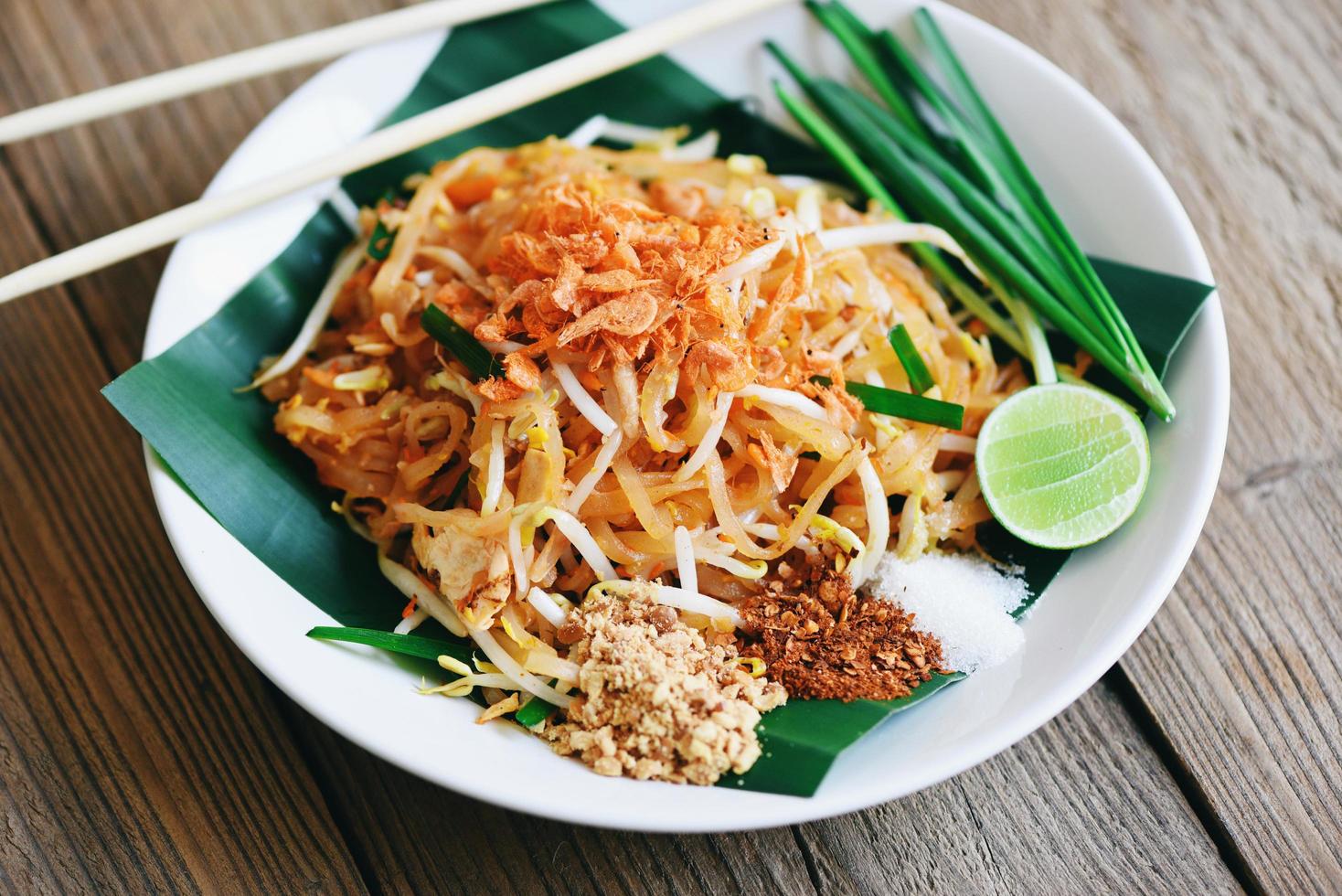 pfannennudelpad thai auf teller serviert auf dem esstisch essen, thailändisches essen gebratene nudeln im thailändischen stil mit garnelen bohnensprossen und garnierung erdnüsse chili puderzucker zitrone limette - draufsicht foto