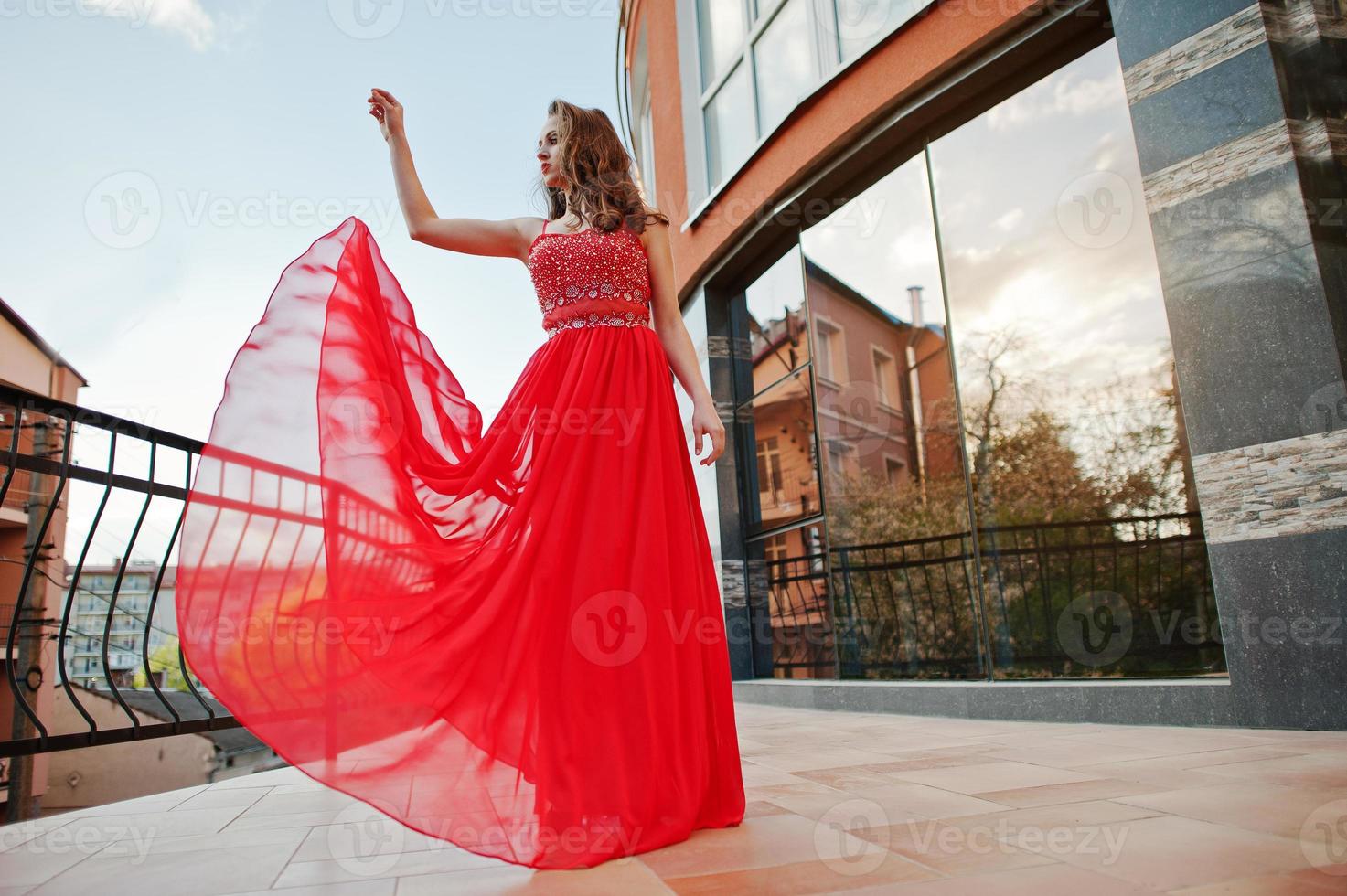 Porträt des modischen Mädchens im roten Abendkleid stellte Hintergrundspiegelfenster des modernen Gebäudes am Terrassenbalkon auf. weht Kleid in die Luft foto