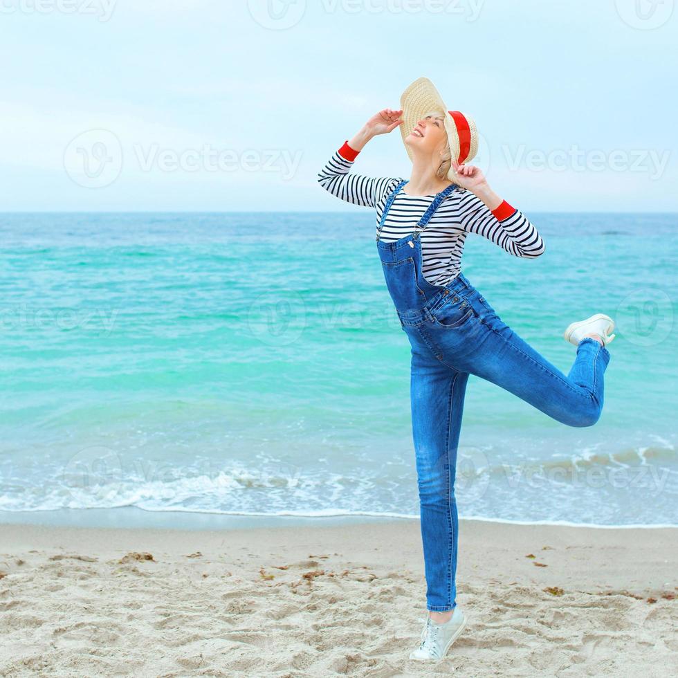Schöne junge blonde kaukasische Frau im Urlaub in gestreifter Bluse, Turnschuhen und Denim-Overall, die am Strand vor dem erstaunlichen blauen Meereshintergrund springt foto