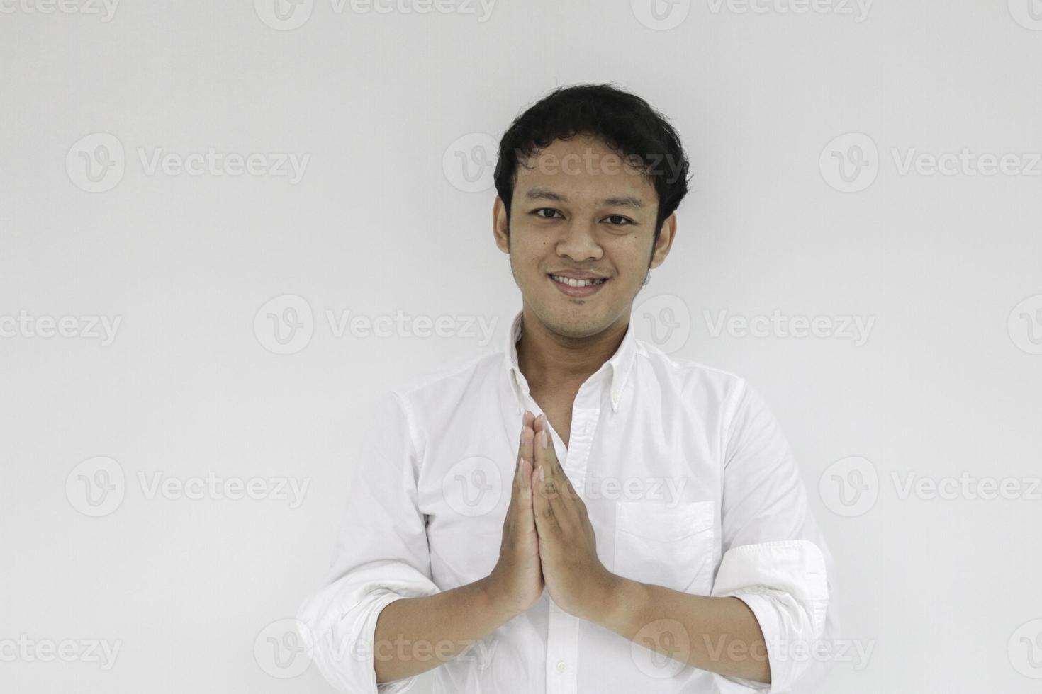 junger asiatischer mann mit weißem hemd gibt grüßende hände mit einem breiten lächeln auf ihrem gesicht. indonesischer Mann auf grauem Hintergrund. Eid Mubarak foto