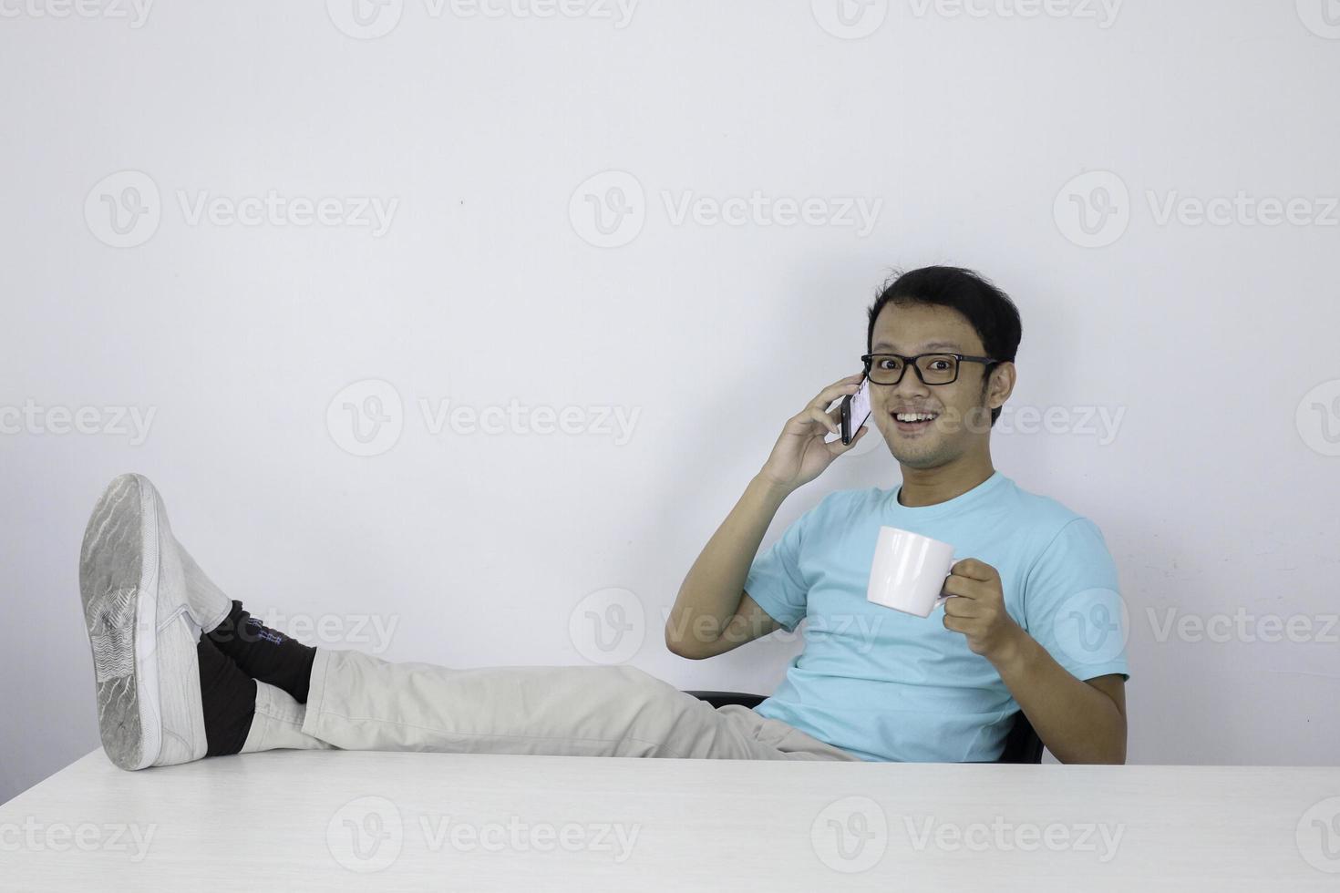 junger asiatischer mann fühlt sich glücklich und lächelt, wenn er mit dem bein auf dem tisch am telefon spricht. foto