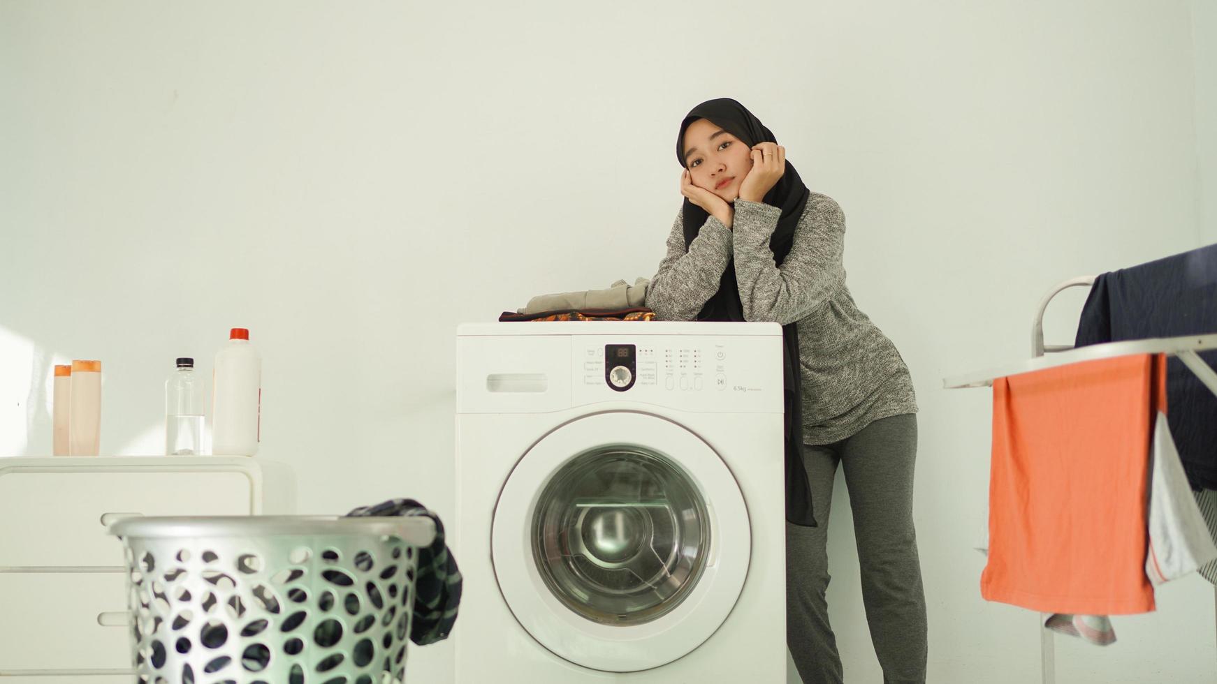 asiatische frau im hijab wartet darauf, dass die waschmaschine zu hause schleudert foto