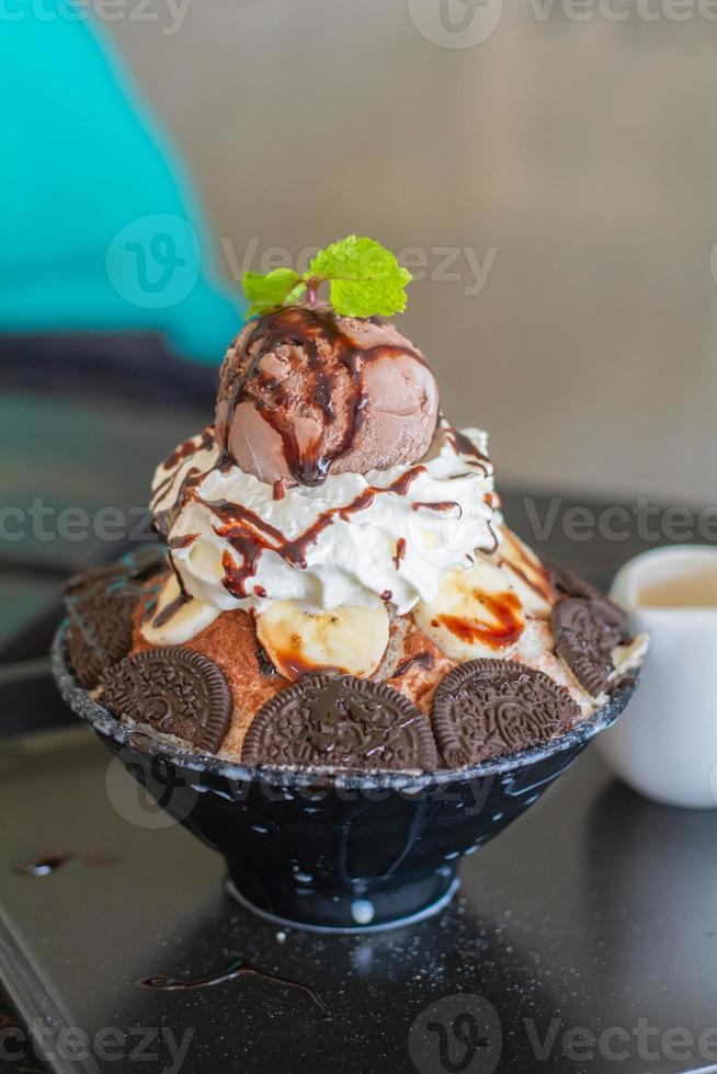 nahaufnahme schokoladeneis, schlagsahne, bananenscheibe auf bingsu in schwarzer schüssel auf holztisch im café und restaurant. leckeres koreanisches rasiertes eis. foto