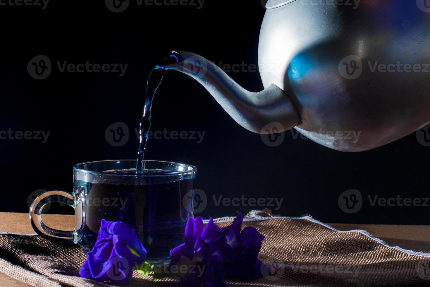 Gießen Sie Schmetterlingserbsentee mit Metalltopf in eine Tasse mit violetter Blume auf braunem Tischtuch auf schwarzem Hintergrund. gesundes getränk zum trinken. kräuter und medizinisches konzept. foto