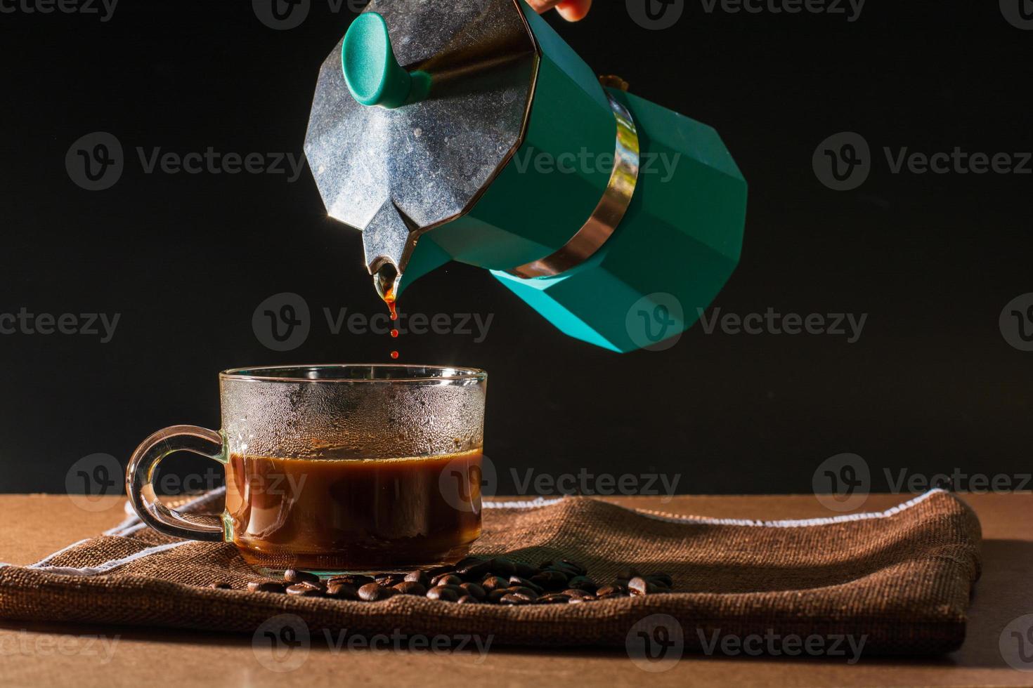 gießen sie heißen schwarzen kaffee aus der grünen moka-kanne in eine klare kaffeetasse mit rauch und kaffeebohnen auf brauner tischdecke und holztisch. nutzen des kaffeekonzepts. foto