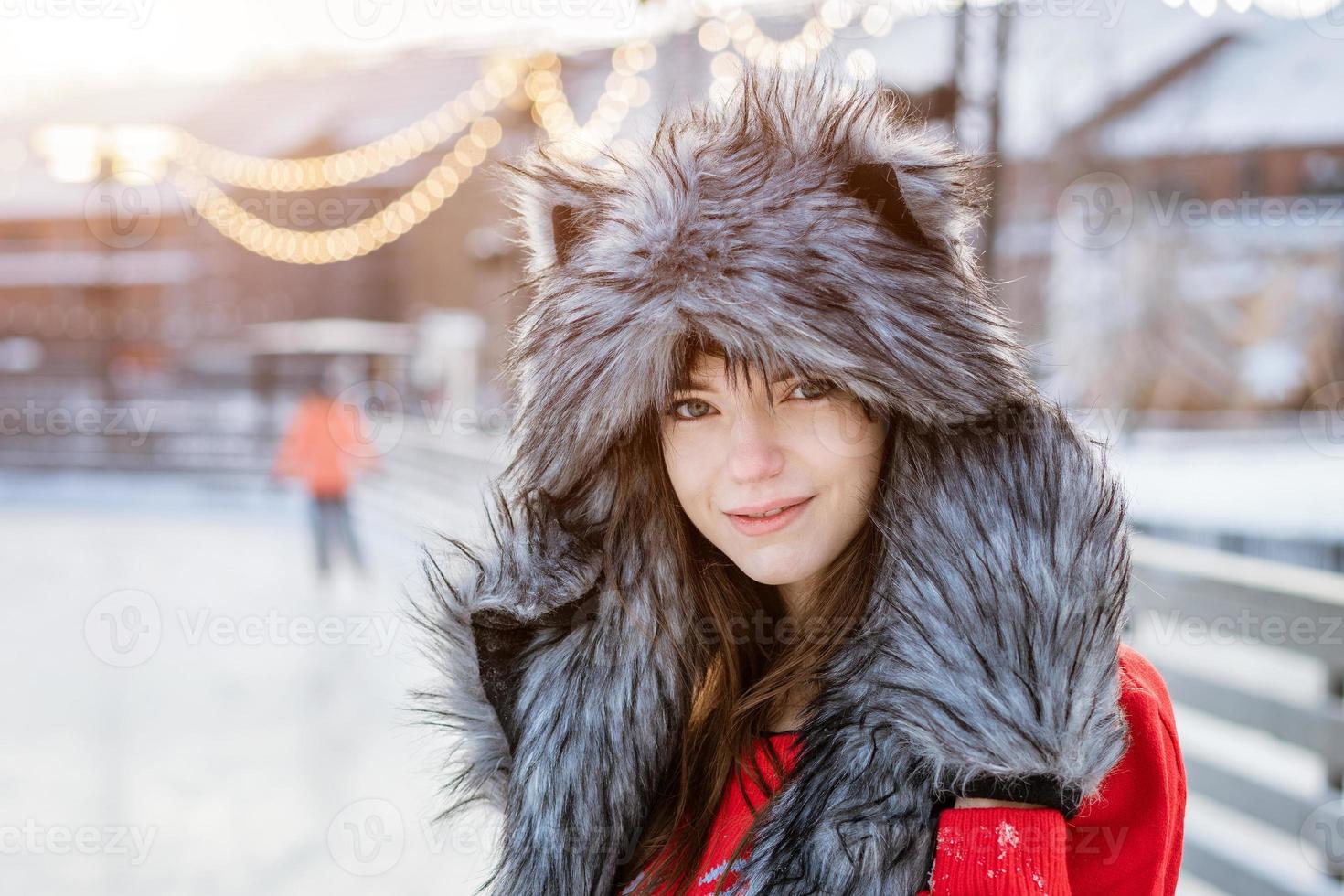 Fröhliche junge Frau mit Wolfshut im Winter auf der Eisbahn, die in einem roten Pullover posiert foto