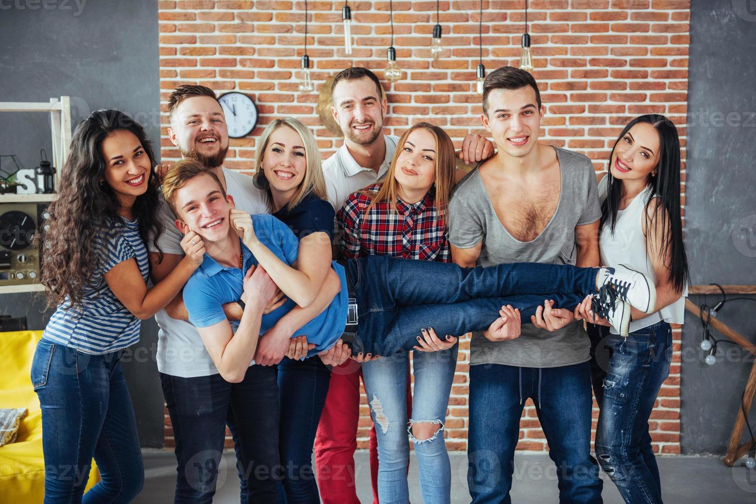 Gruppenporträt von multiethnischen Jungen und Mädchen mit farbenfrohen, modischen Kleidern, die einen Freund halten, der auf einer Ziegelwand posiert, Menschen im urbanen Stil, die Spaß haben, Konzepte über den Lebensstil der Jugendzusammengehörigkeit foto