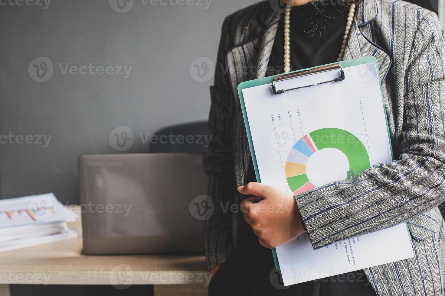 Abgeschnittene Aufnahme einer Geschäftsfrau, die in einem modernen Büro mit grauem Hintergrund ein Donut-Diagramm in der Zwischenablage hält. Vorbereitung auf Meetings oder Präsentationen foto