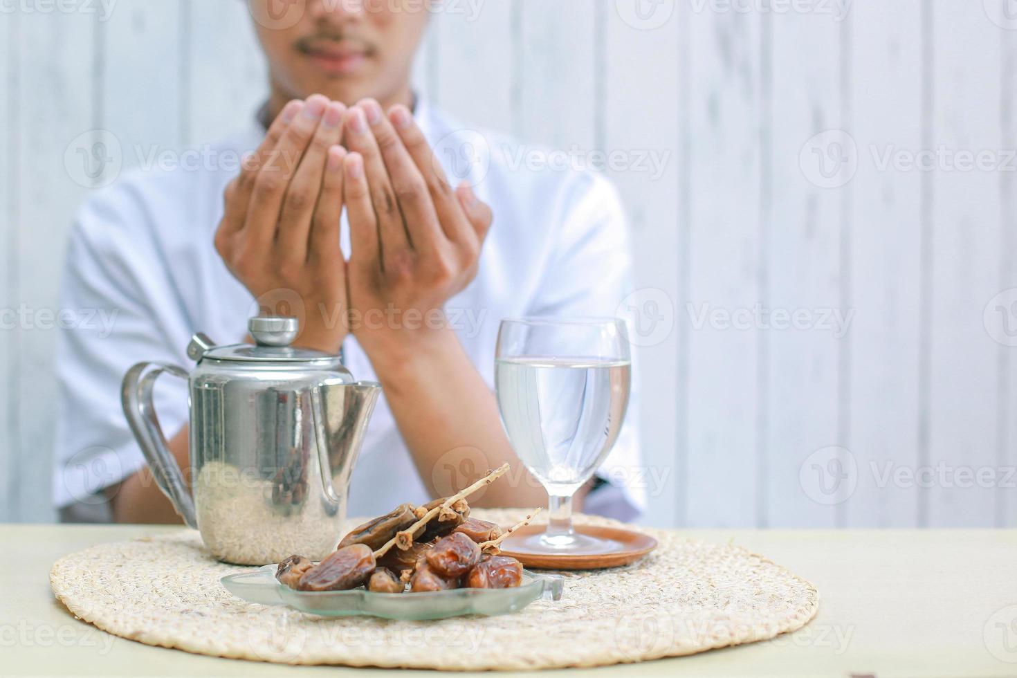 Iftar-Gericht mit muslimischer Mannhand, die zu Allah betet. datteln obst mit glas mineralwasser und teekanne auf dem tisch. traditionelles ramadan, iftar-mahlzeit. ramadan kareem fastenmonatskonzept. foto