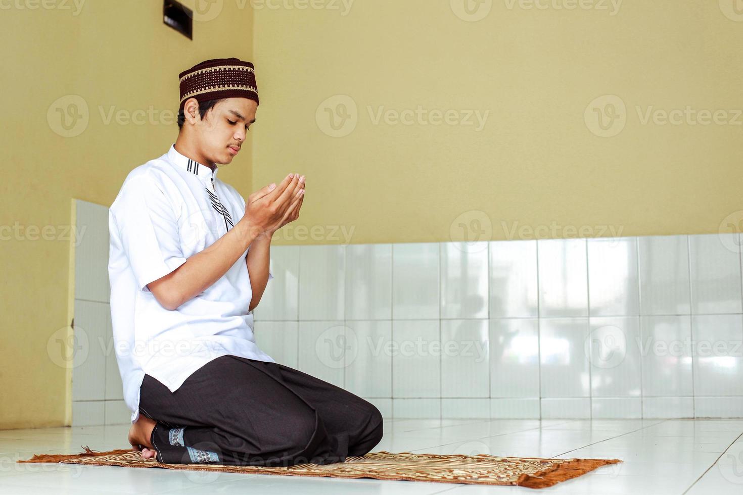 seitenansicht des jungen asiatischen muslims, der zu allah betet. islamkerl, der mit bettelnder handbewegung auf der gebetsmatte sitzt foto