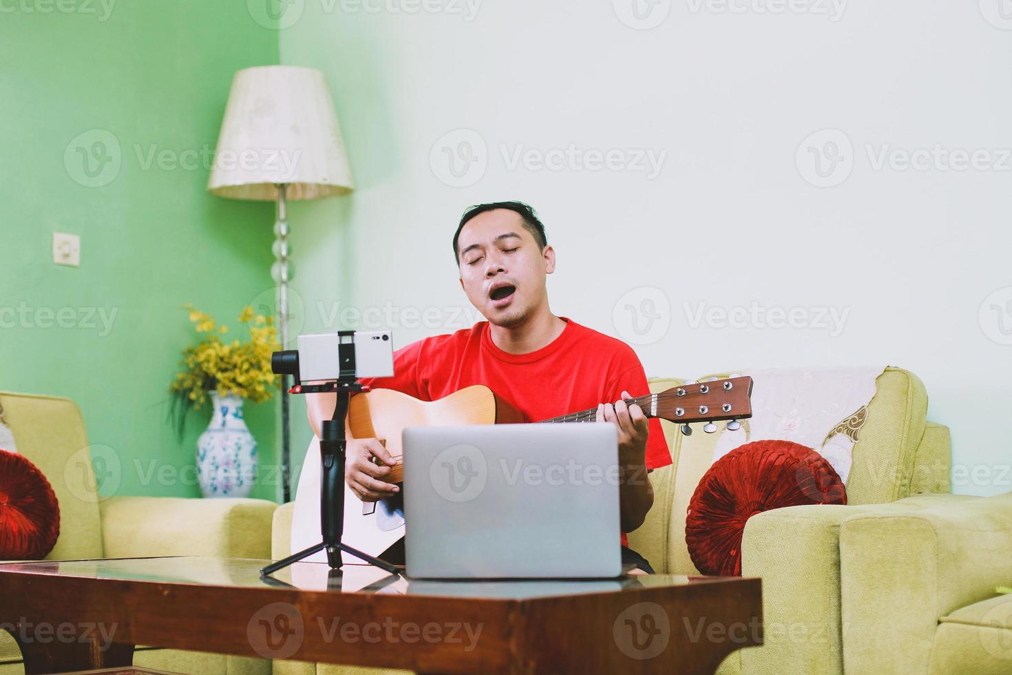 Junger Mann singt und spielt Gitarre, während er zu Hause per Smartphone und Laptop aufnimmt foto