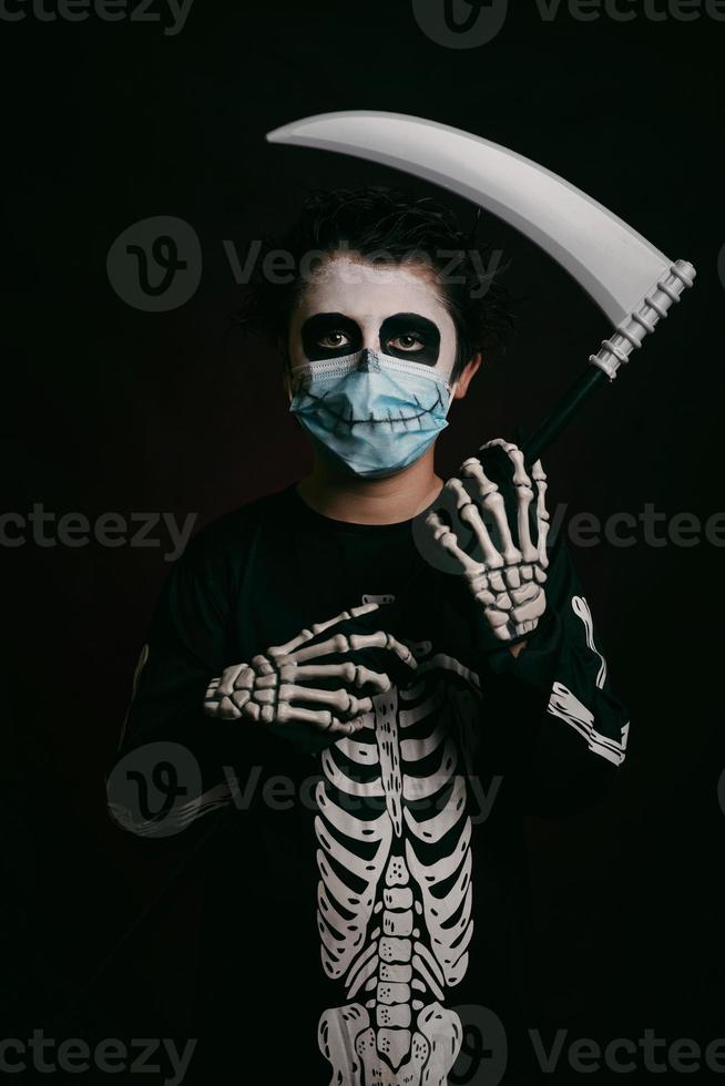 fröhliches halloween, kind mit medizinischer maske in einem skelettkostüm mit einer sense foto