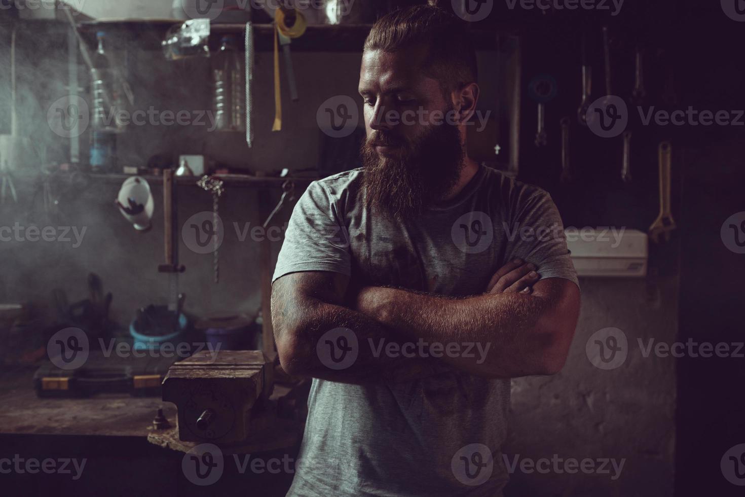 Schöner brutaler Mann mit Bart, der in seiner Garage vor dem Hintergrund von Reparaturwerkzeugen steht foto