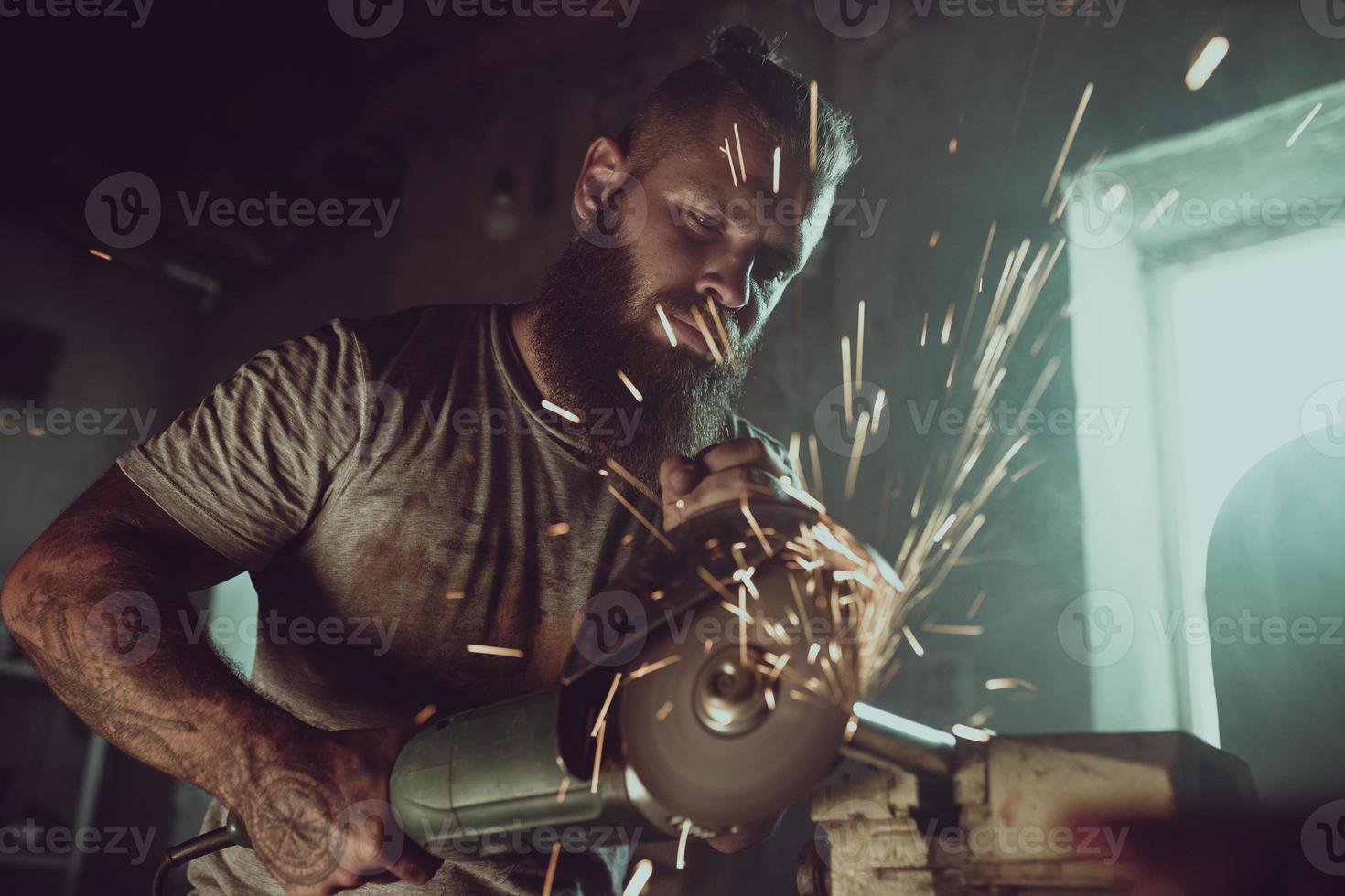 Schöner brutaler Mann mit Bart, der ein Motorrad in seiner Garage repariert und mit einer Kreissäge arbeitet. in der garage viele funken und rauch vom sägen foto