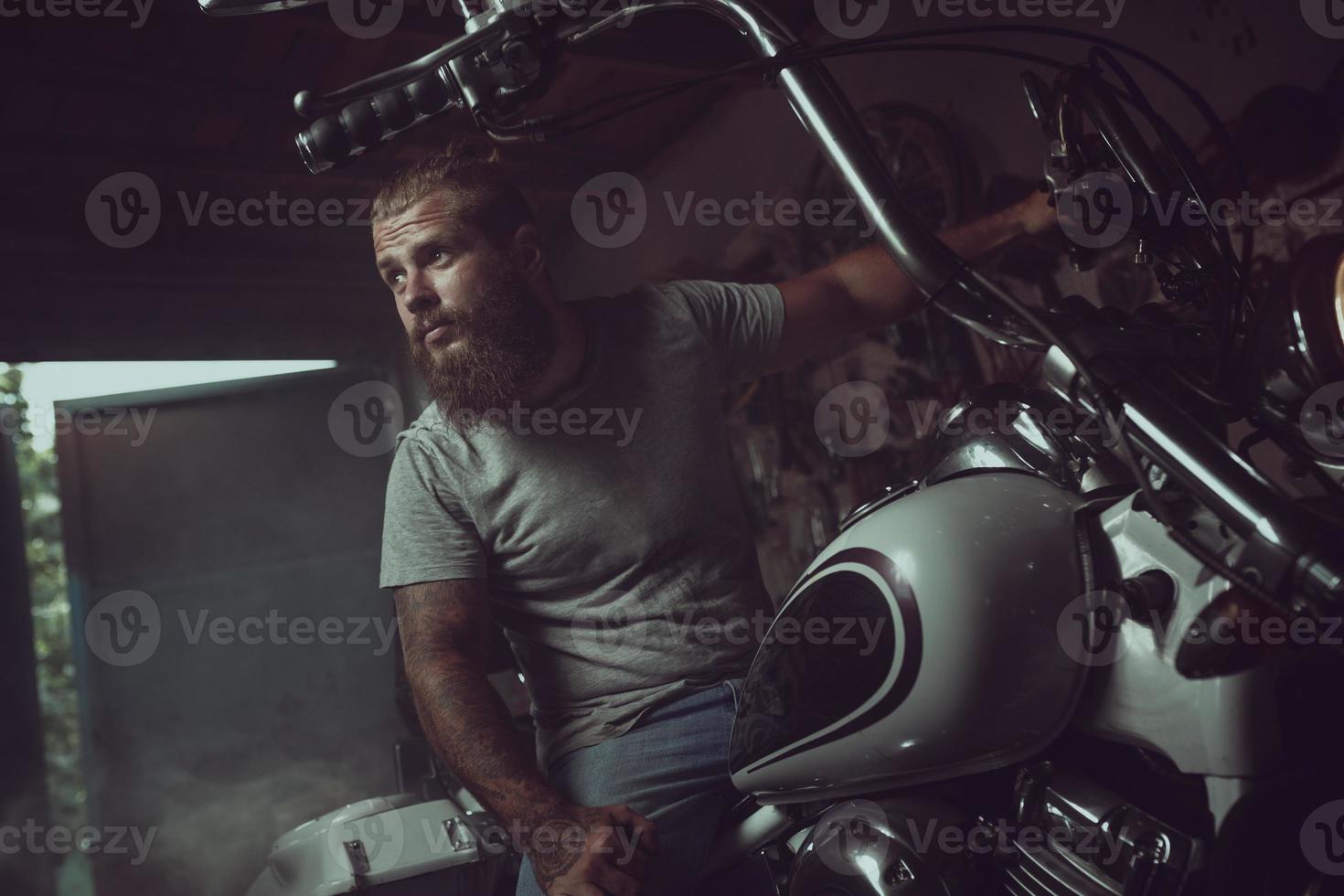 Schöner brutaler Mann mit Bart, der in seiner Garage auf einem Motorrad sitzt und wegschaut foto