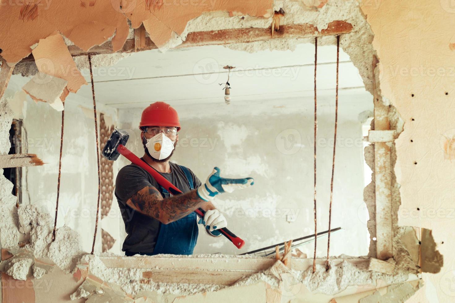 baumeister mit einem hammer in den händen bricht die zementwand der baumeister trägt einen schutzanzug und einen helm foto