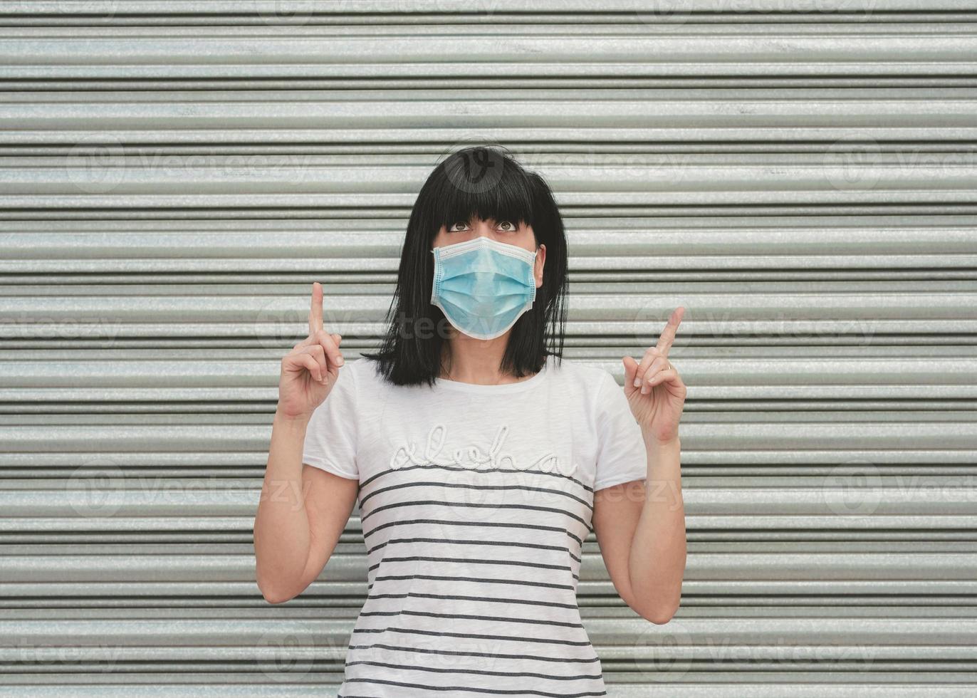 frau, die eine medizinische maske trägt, die nachdenklich mit zwei zeigefingern nach oben zeigt und nach oben schaut foto