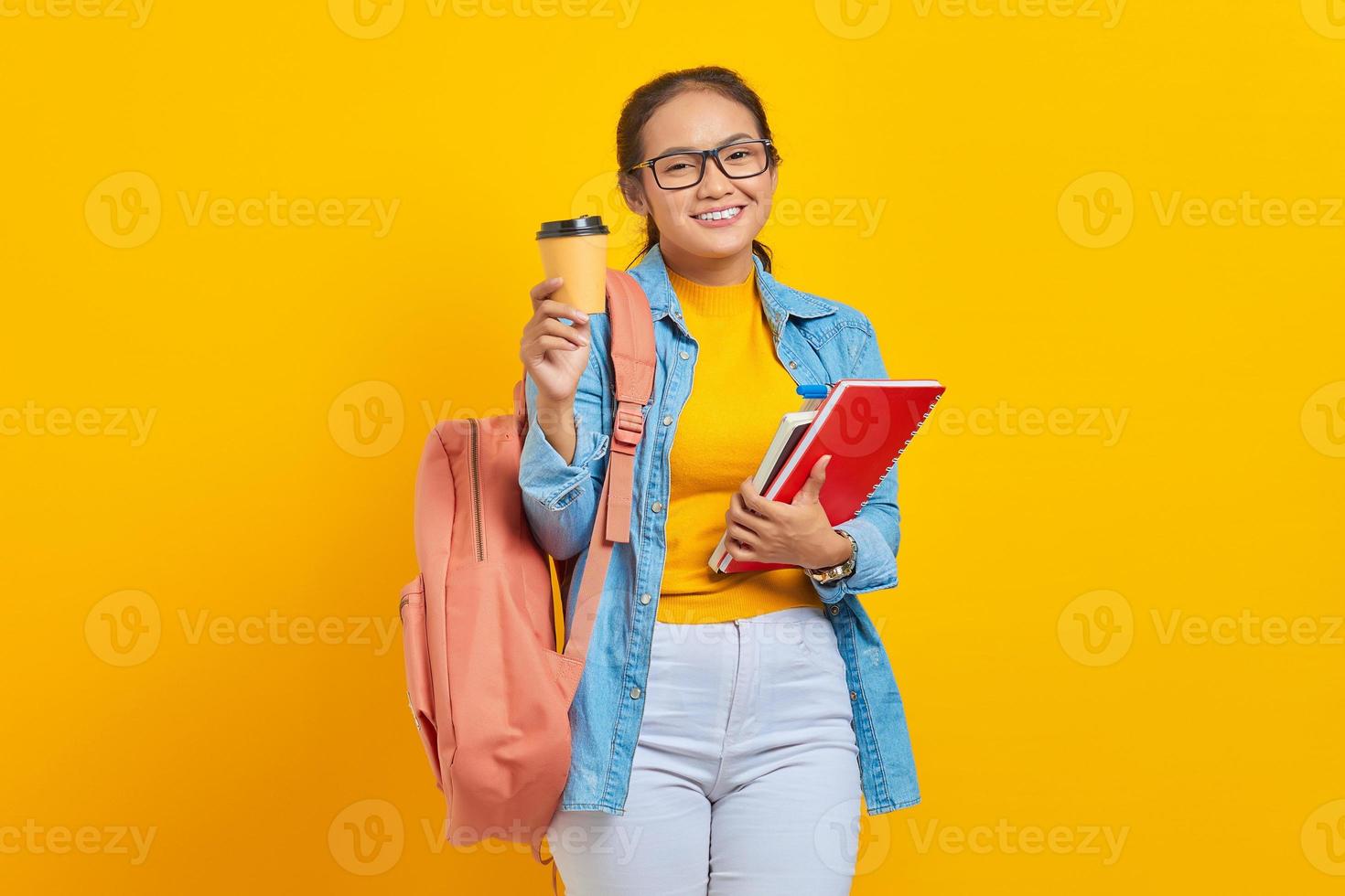 porträt einer fröhlichen jungen asiatischen studentin in lässiger kleidung mit rucksack, der ein buch hält und kaffee zum mitnehmen zeigt, der die kamera isoliert auf gelbem hintergrund betrachtet foto