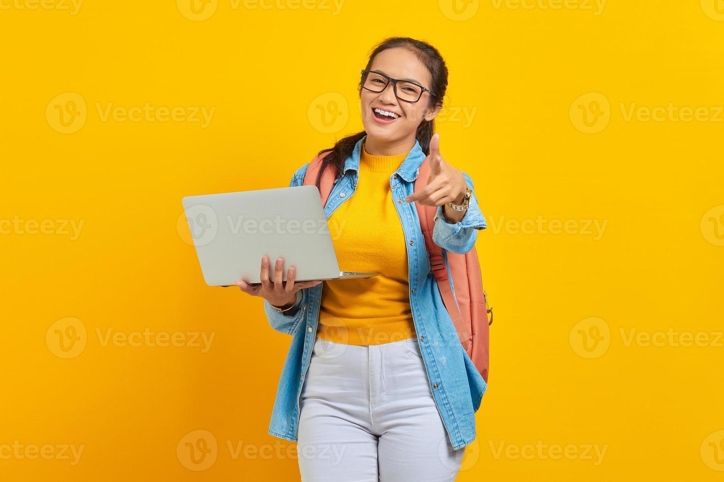 porträt einer fröhlichen jungen asiatischen studentin in lässiger kleidung mit rucksack, die einen laptop benutzt und mit dem finger isoliert auf gelbem hintergrund auf die kamera zeigt. Bildung im College-Universitätskonzept foto