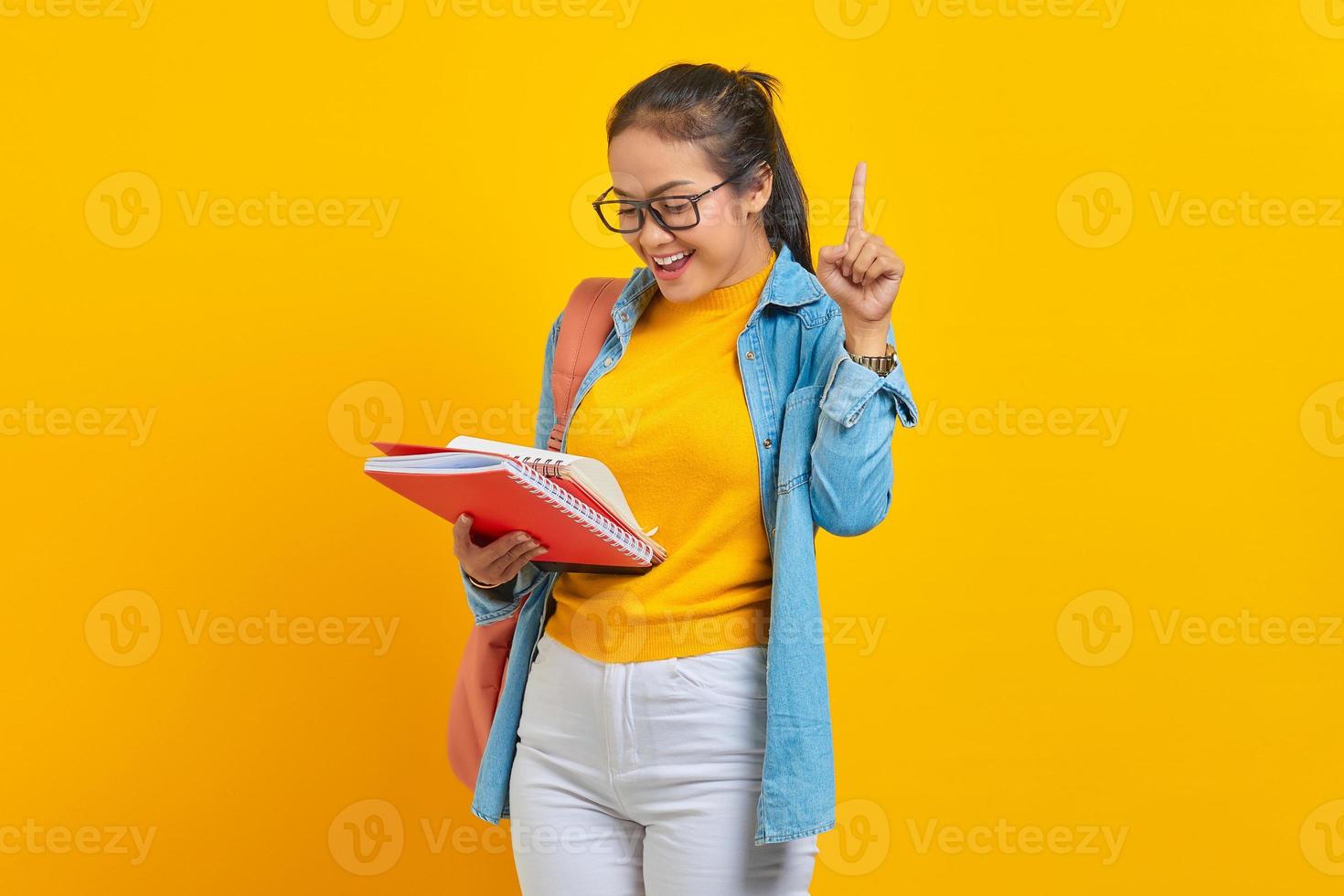 Fröhliche junge Studentin in Denim-Kleidung mit Rucksack, Buch mit dem Zeigefinger nach oben mit toller neuer Idee auf gelbem Hintergrund. Bildung im High School University College-Konzept foto