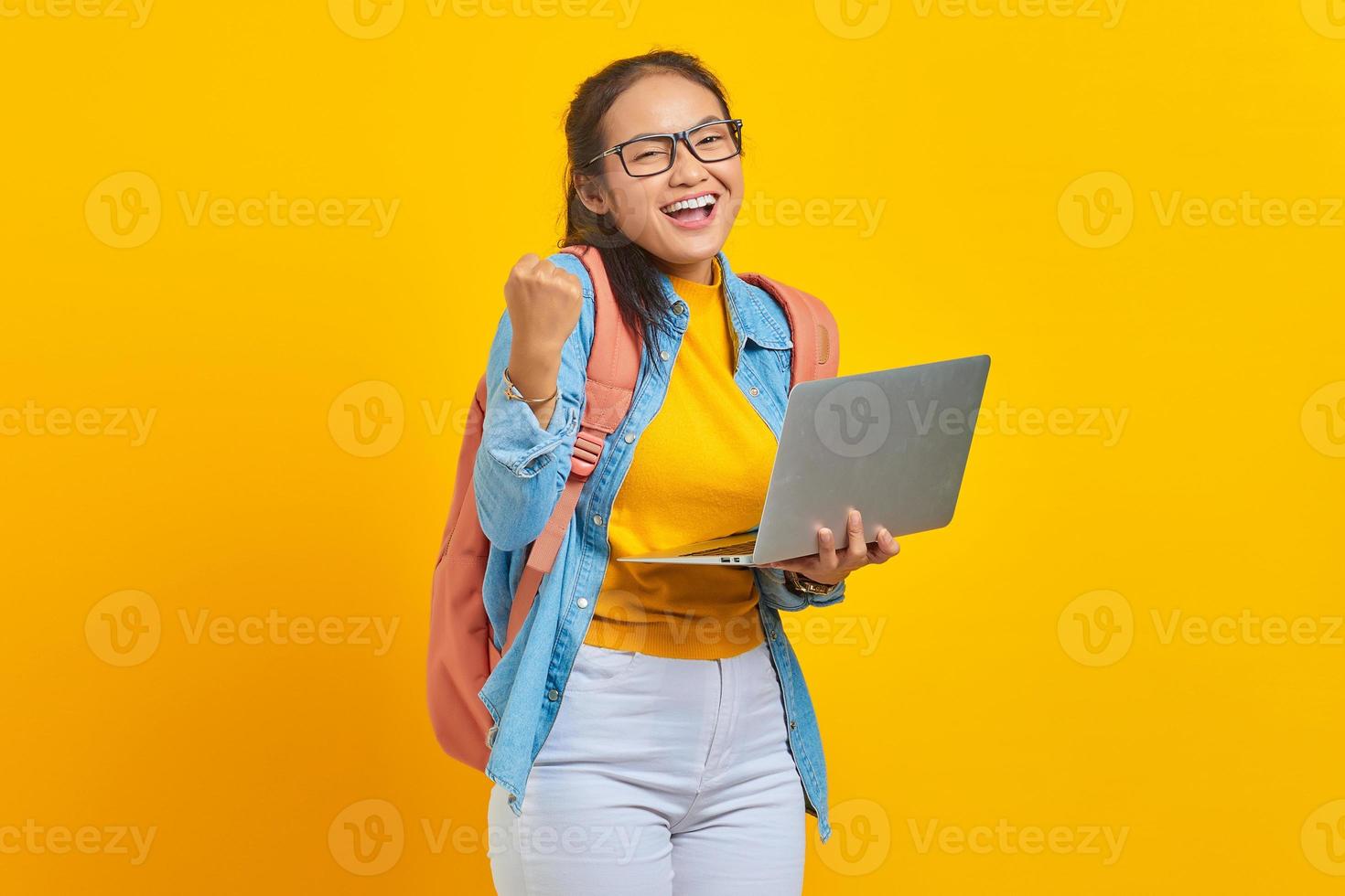 porträt einer aufgeregten jungen asiatischen studentin in lässiger kleidung mit rucksack, die einen laptop benutzt und den erfolg isoliert auf gelbem hintergrund feiert. ausbildung im universitätshochschulkonzept foto