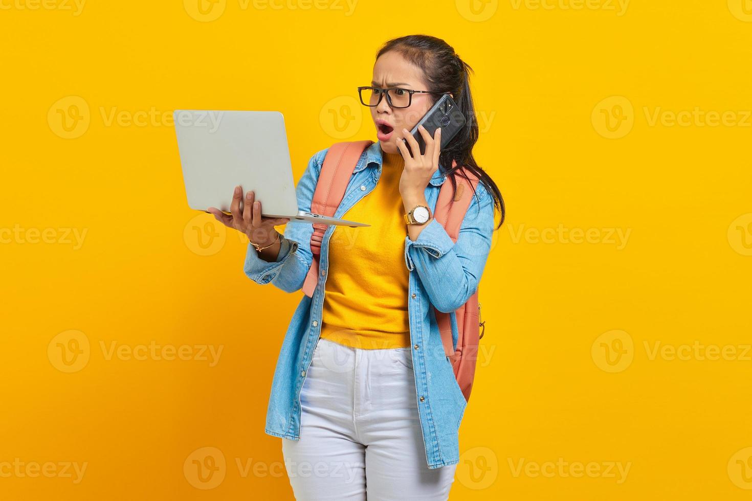 porträt einer überraschten jungen asiatischen studentin in lässiger kleidung mit rucksack, die am handy spricht und einen laptop isoliert auf gelbem hintergrund verwendet. Bildung im College-Universitätskonzept foto