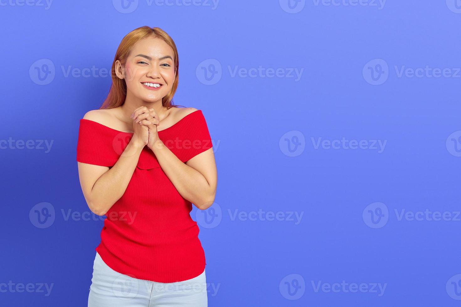 Lächelnde schöne asiatische Frau in rotem Kleid, die sich die Hände reibt und selbstbewusst isoliert auf violettem Hintergrund aussieht foto