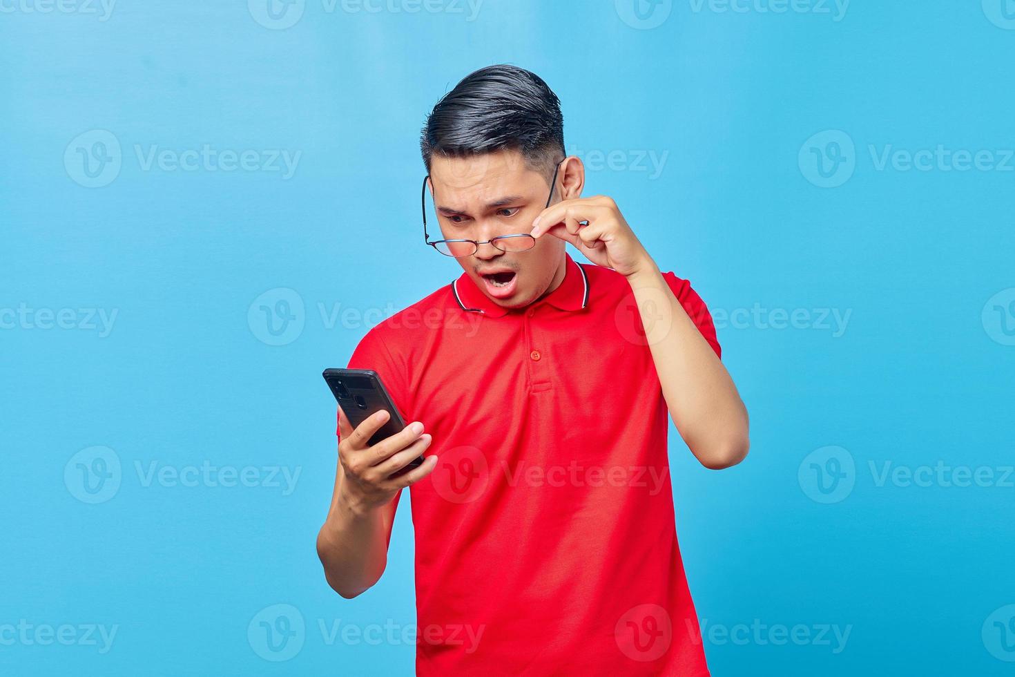 Porträt eines überraschten asiatischen jungen Mannes, der eine Brille hält und eingehende Nachrichten auf dem Smartphone liest, isoliert auf blauem Hintergrund foto