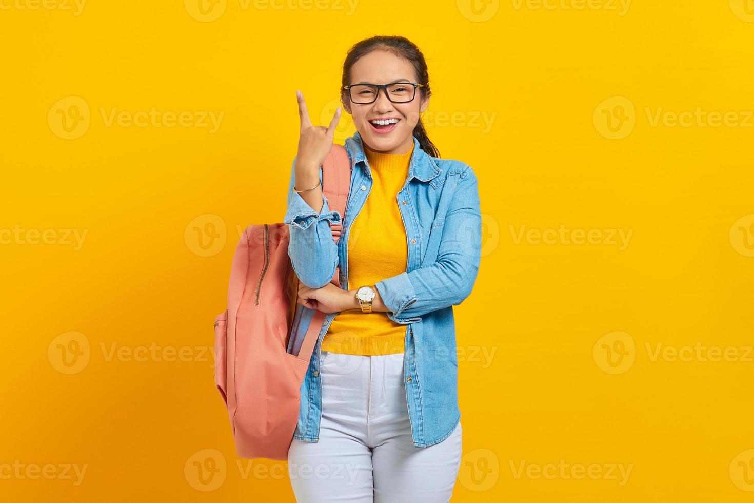 Schöne junge asiatische Studentin im Denim-Outfit mit Rucksack, die mit verrücktem Ausdruck schreit und Rocksymbol mit erhobenen Händen isoliert auf gelbem Hintergrund macht. ausbildung im universitätshochschulkonzept foto