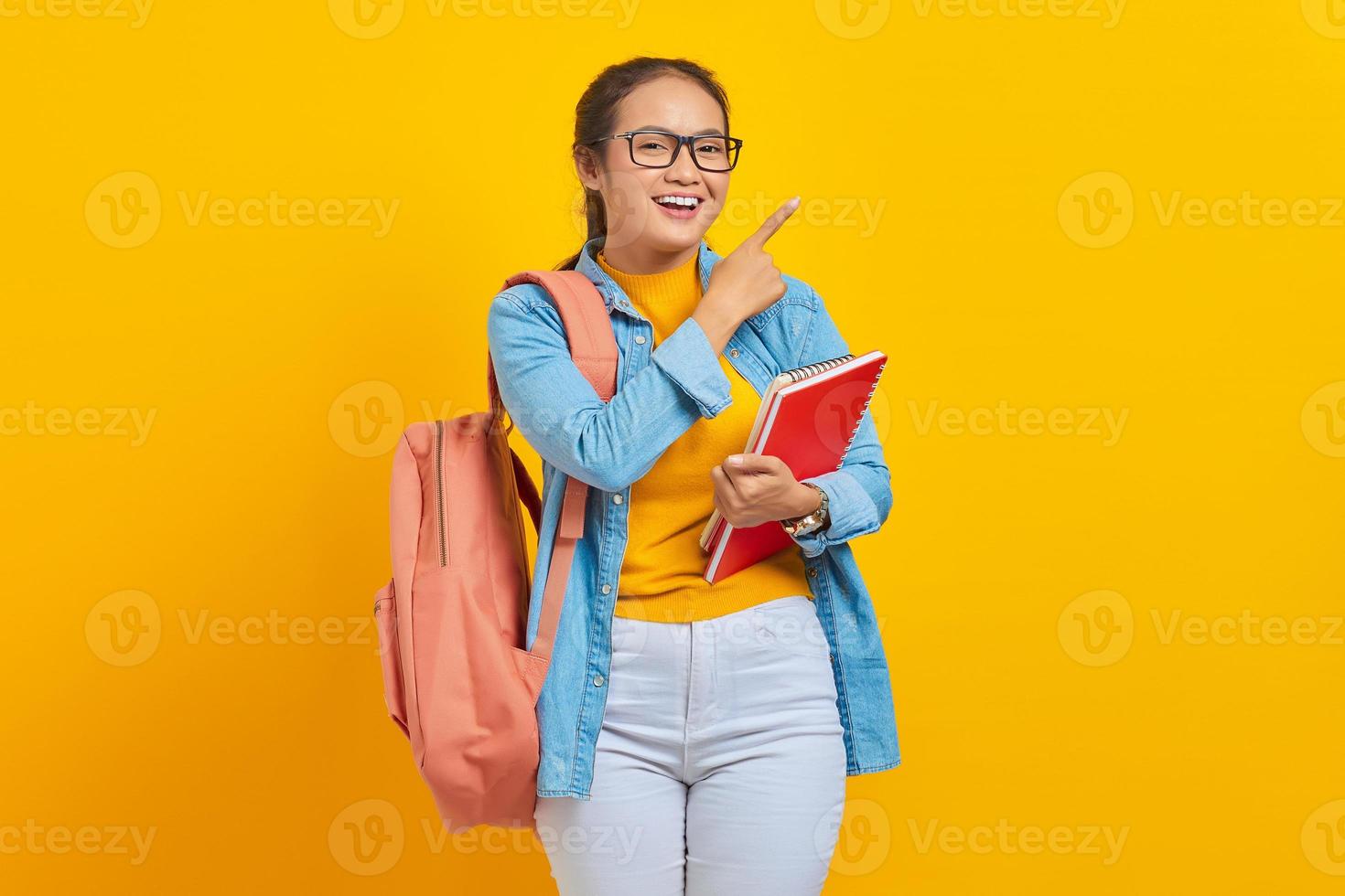 Porträt einer lächelnden jungen asiatischen Studentin in Freizeitkleidung mit Rucksack, der ein Buch hält und mit dem Finger isoliert auf gelbem Hintergrund zur Seite zeigt. Bildung im College-Universitätskonzept foto