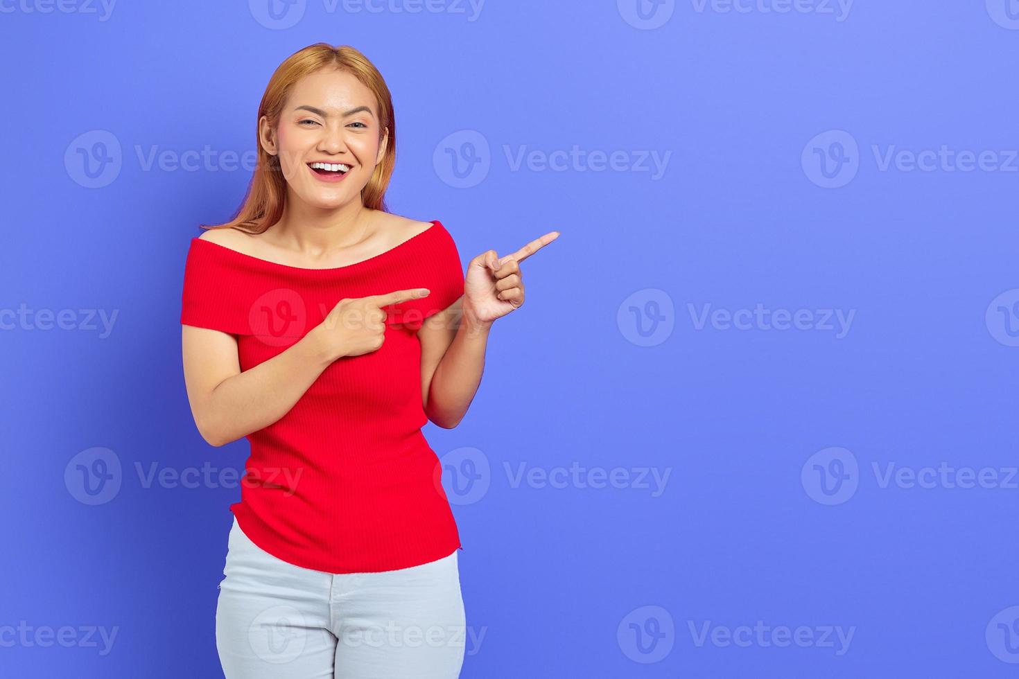 porträt einer fröhlichen jungen asiatischen frau, die steht und zeigt, um raum mit dem finger zu kopieren, der auf violettem hintergrund isoliert ist foto