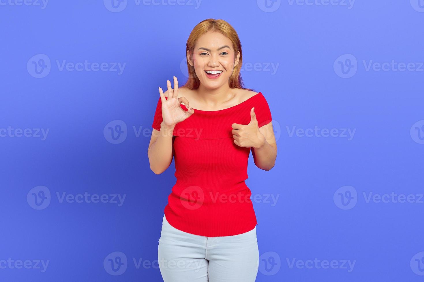 Porträt einer schönen, fröhlichen asiatischen Frau, die ein rotes Kleid trägt und lächelt, während sie mit dem Finger auf violettem Hintergrund ein Ok-Zeichen und eine Geste der Zustimmung zeigt foto