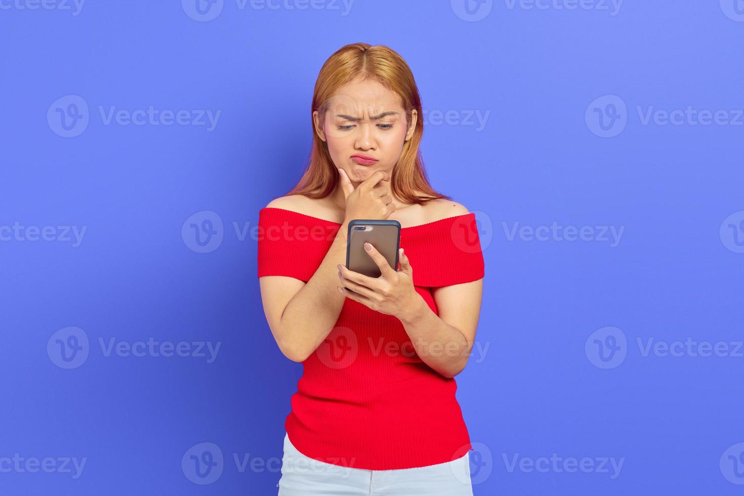 Porträt einer unzufriedenen jungen asiatischen Frau, die die Stirn runzelt, während sie das Smartphone isoliert auf violettem Hintergrund hält foto