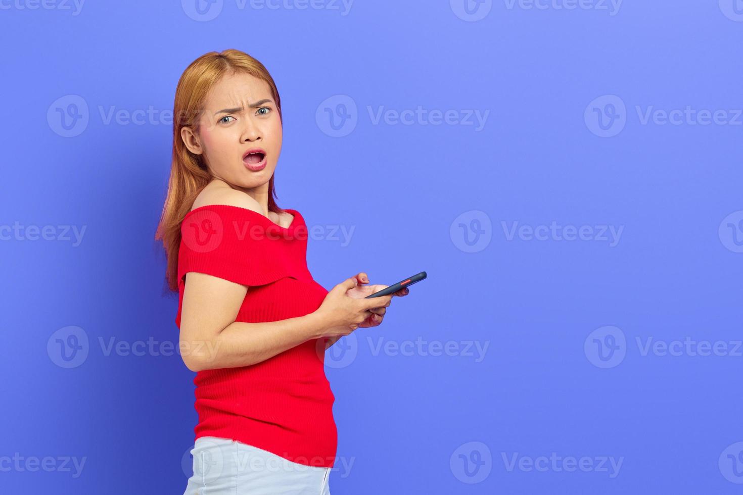 Porträt einer überraschten jungen asiatischen Frau in rotem Kleid, kurzes blondes Haar mit Handy isoliert auf violettem Hintergrund foto