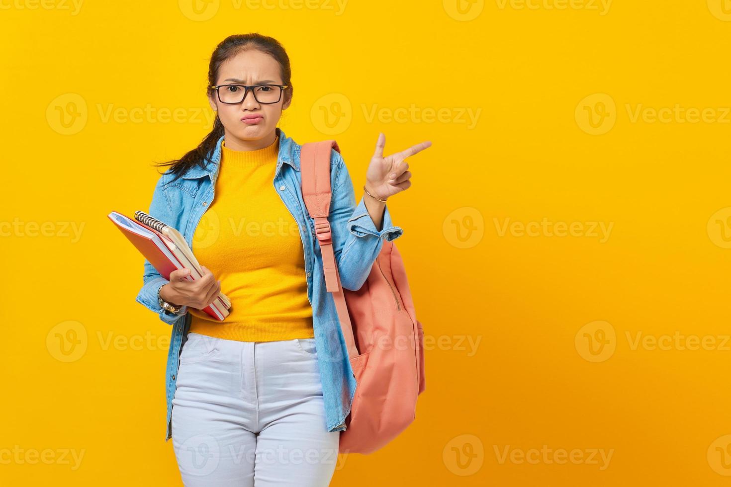 porträt einer verwirrten jungen asiatischen studentin in jeanskleidung mit rucksack, der notizbuch hält und mit dem finger auf den kopierraum zeigt, der auf gelbem hintergrund isoliert ist foto