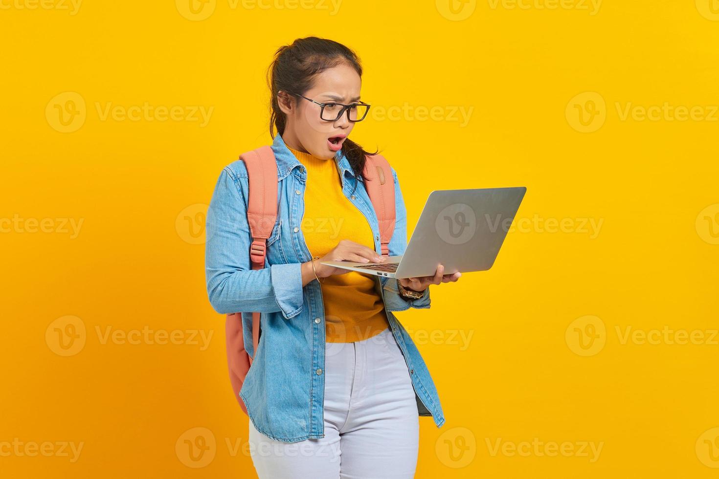 Porträt einer überraschten jungen asiatischen Studentin in Freizeitkleidung mit Rucksack, die eingehende E-Mails auf einem Laptop isoliert auf gelbem Hintergrund betrachtet. ausbildung im universitätshochschulkonzept foto