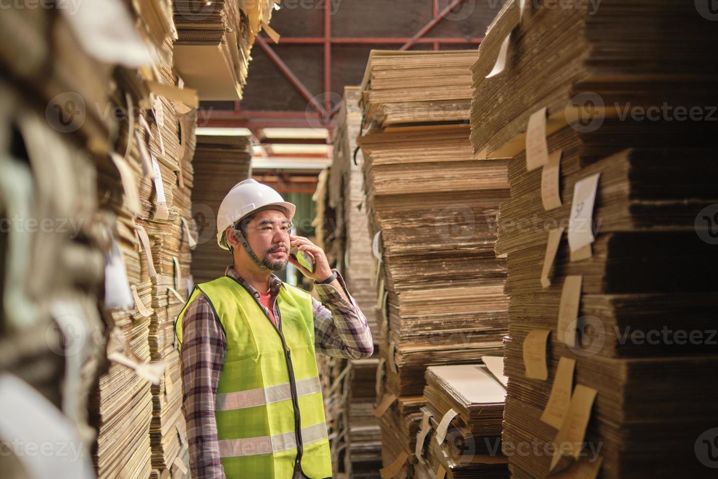 asiatischer männlicher arbeiter in sicherheitsuniform und helm, vorgesetzter inspektor spricht mit dem handy über die verpackungslagerbestellung in der kistenfabrik mit stapeln von papierstapeln, recyclingproduktionsindustrie. foto