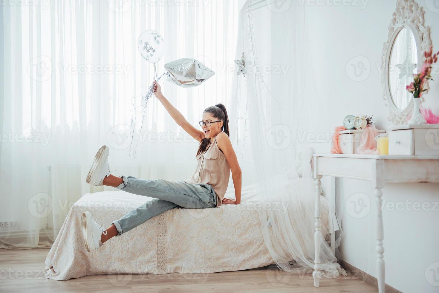 Nahaufnahme süßes brünettes Mädchen im Studio, breit lächelnd und mit transparenten und silbernen Luftballons spielend. Sie trägt eine Brille und verdrehtes Haar. foto